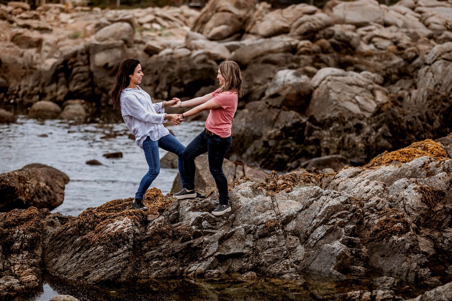 Two women hold hands and balance together on seaside rocks near the ocean.