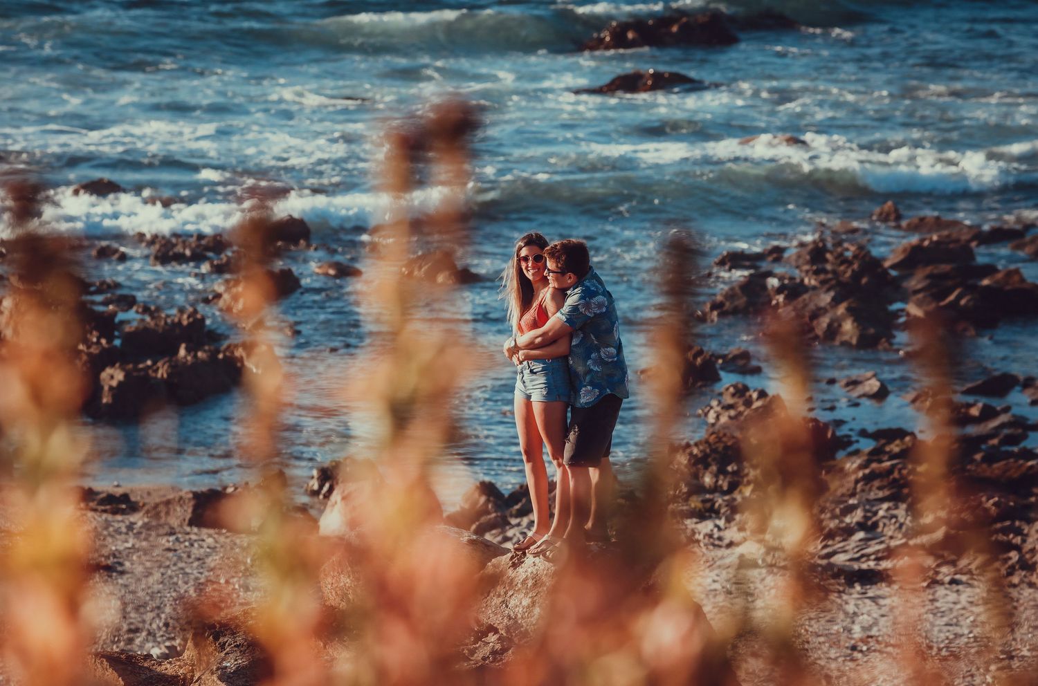 Couple embracing on rocky beach framed by golden coastal wildflowers.