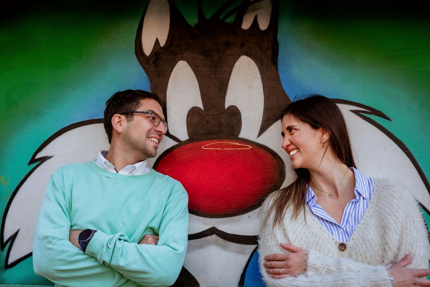 Smiling couple posing in front of colorful Sylvester the Cat cartoon mural looking at each other.