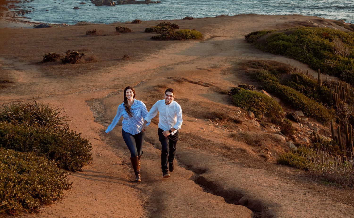 Couple running together on a coastal dirt path with ocean in background.