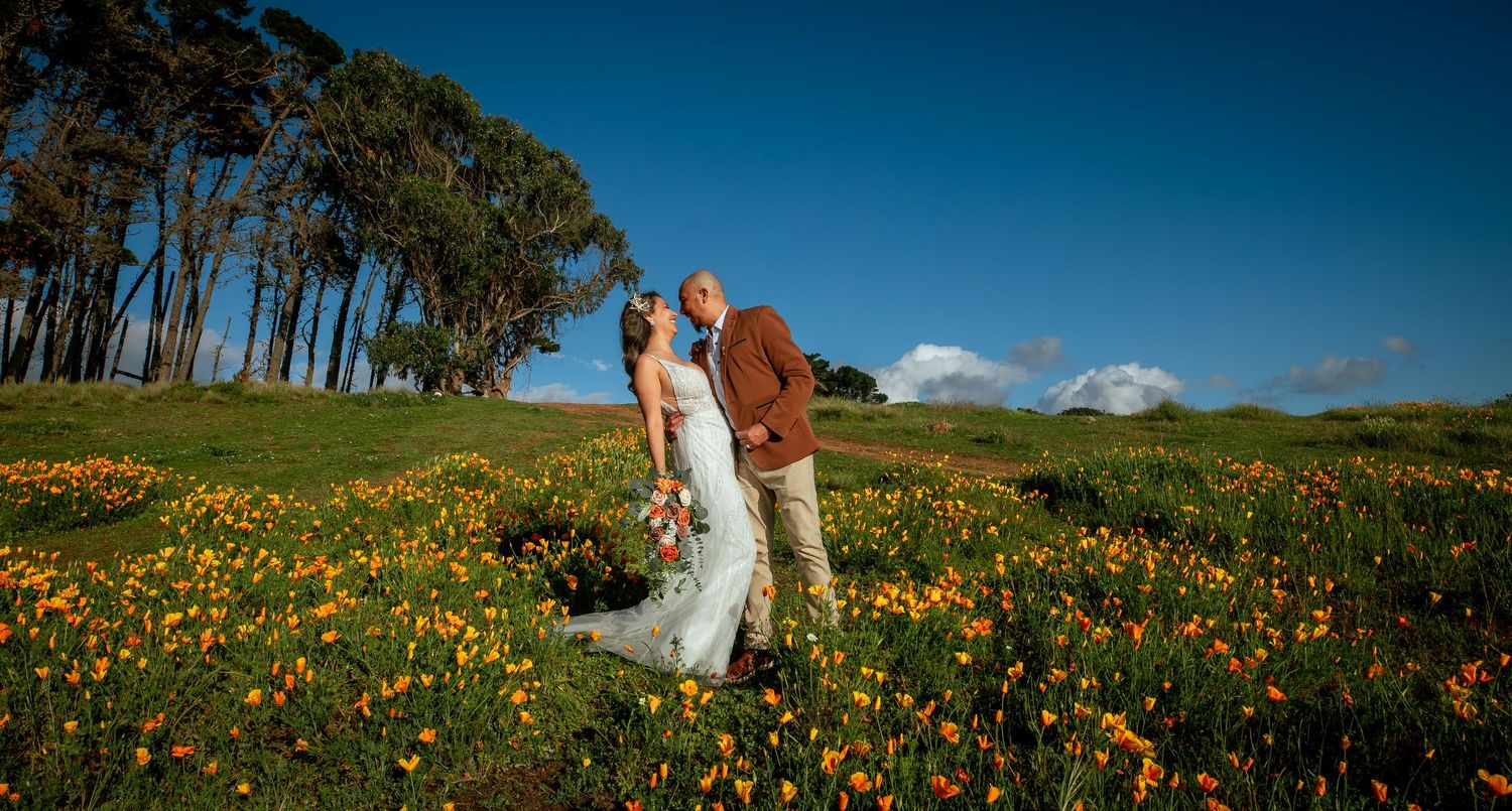 Bride in a white gown and groom in a brown jacket share a kiss in a field of orange wildflowers beneath tall trees and a blue sky.