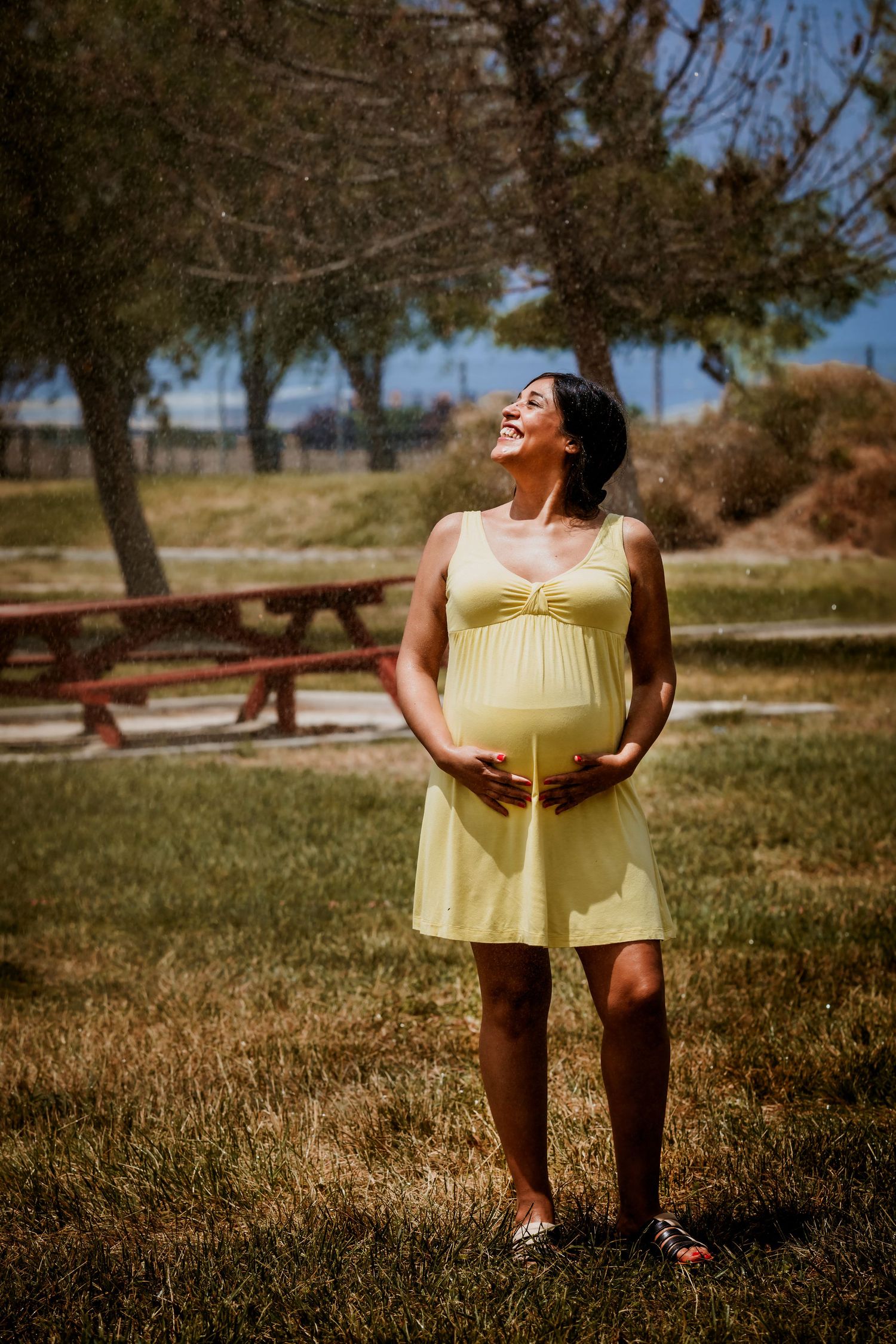 Smiling pregnant woman in yellow dress cradling baby bump outdoors in sunny park setting.
