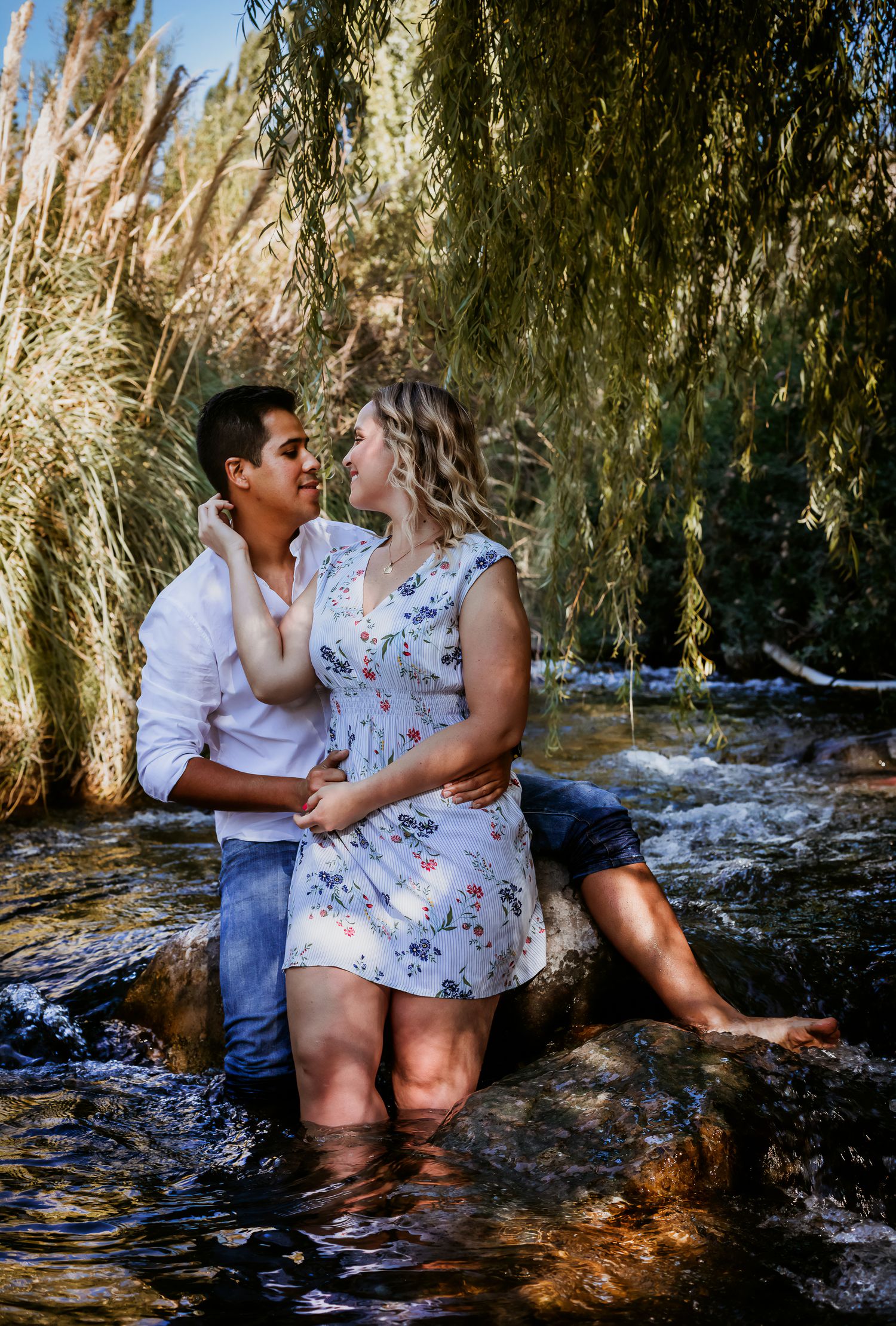 Couple standing in a stream surrounded by lush greenery, sharing a romantic moment on a sunny day.