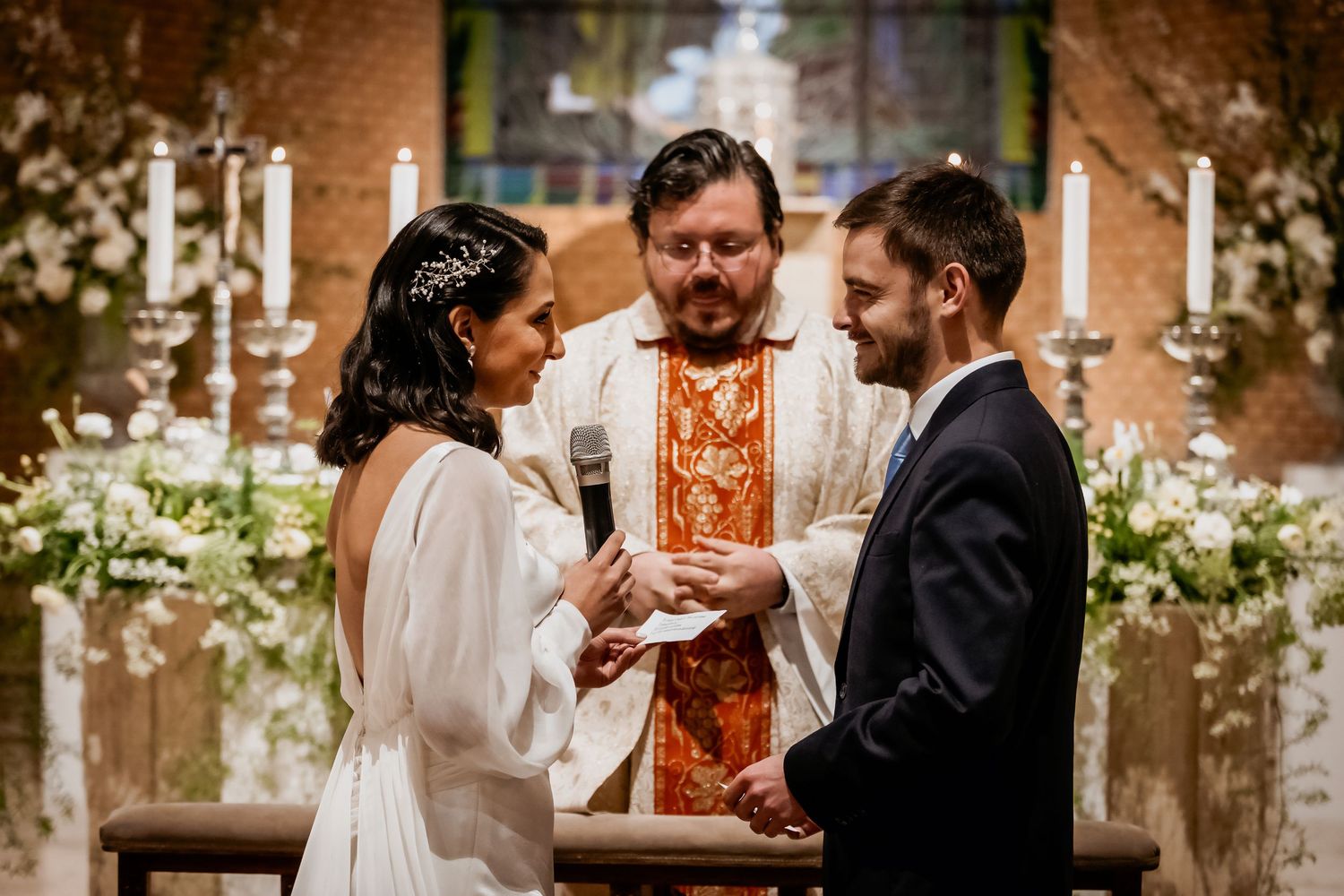 Bride and groom exchange rings at altar with officiant during elegant indoor wedding ceremony with candles and white flowers.
