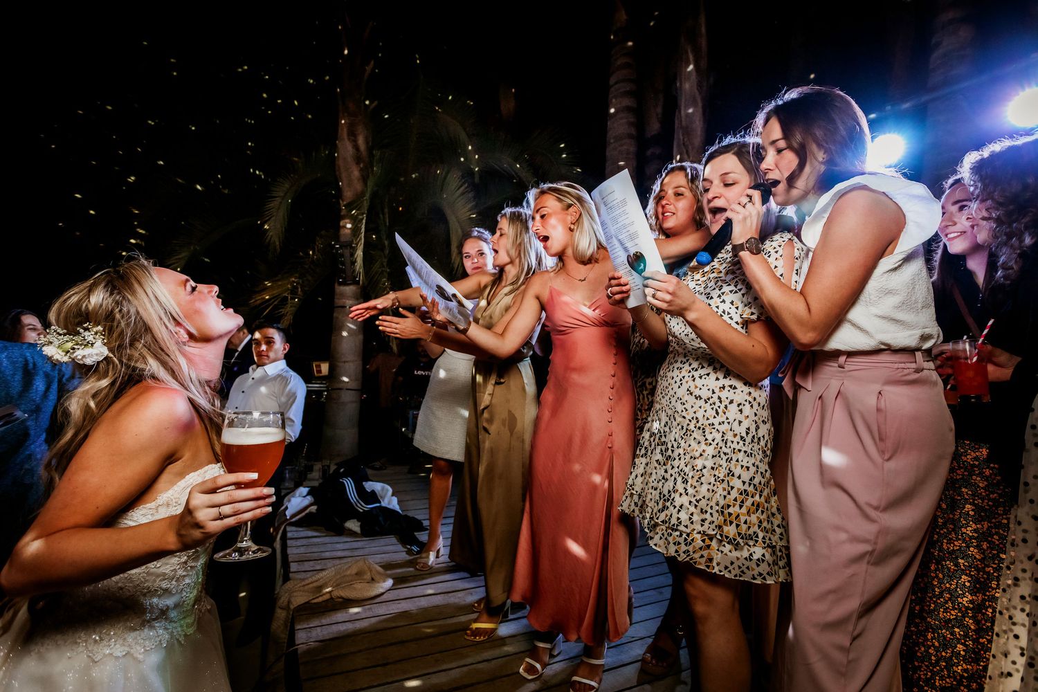 Bride and bridesmaids celebrating with confetti at outdoor evening wedding reception under string lights.