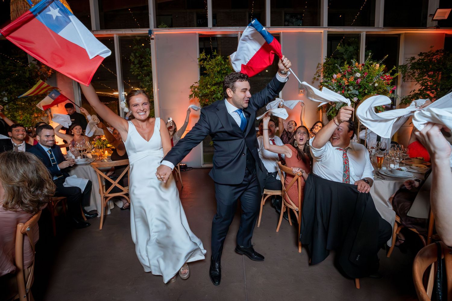 Bride and groom entering wedding reception waving Chilean flags while guests cheer and celebrate joyfully.