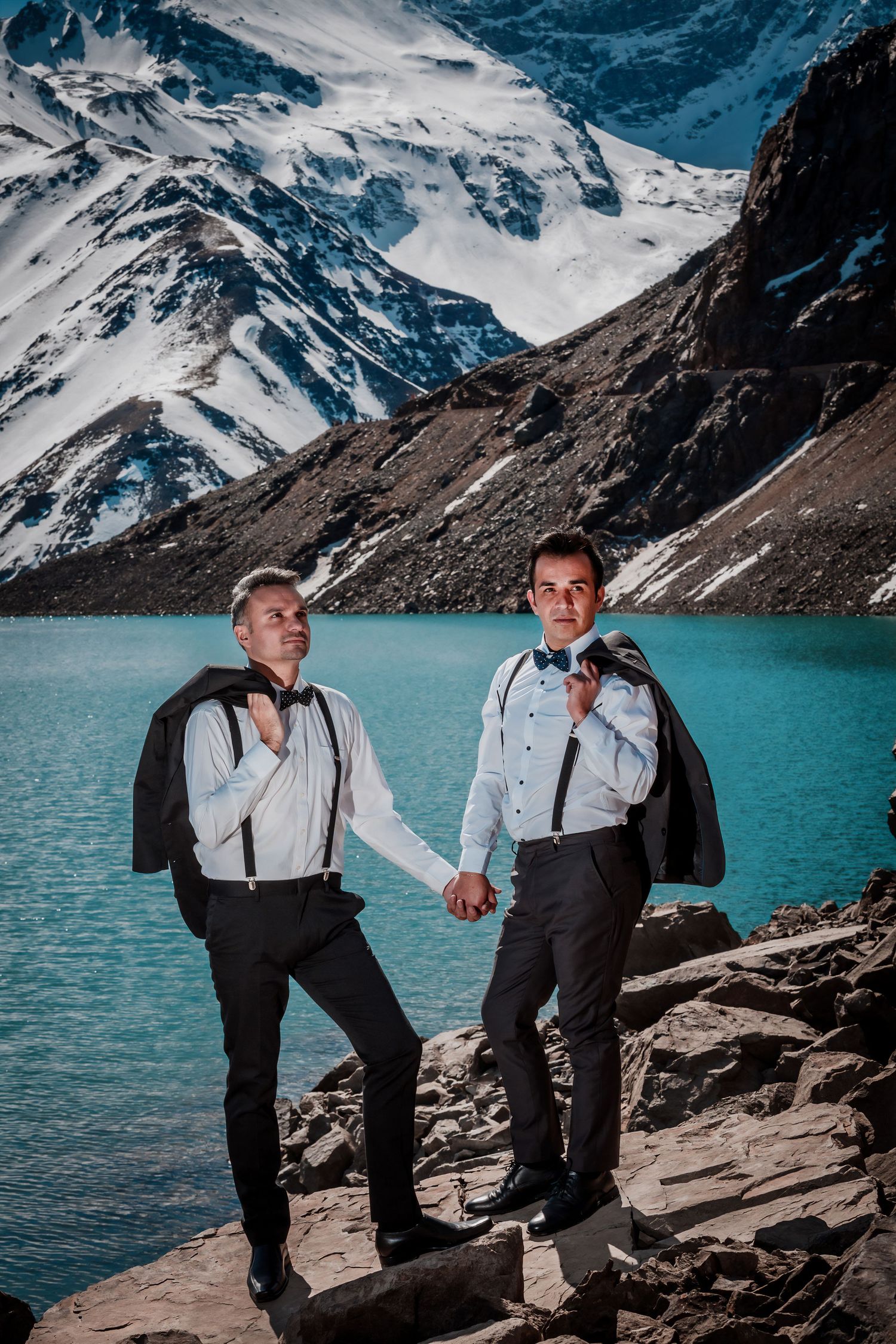 Same-sex couple in tuxedos holding hands by a vivid turquoise alpine lake with snowy mountains.