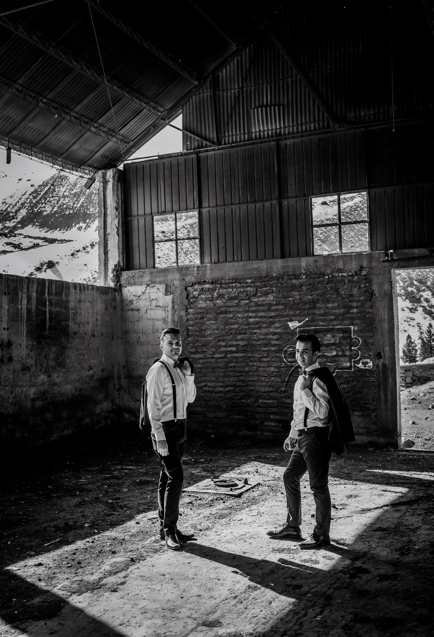 Black and white photo of two grooms in suspenders standing inside a rustic abandoned barn with dramatic lighting.