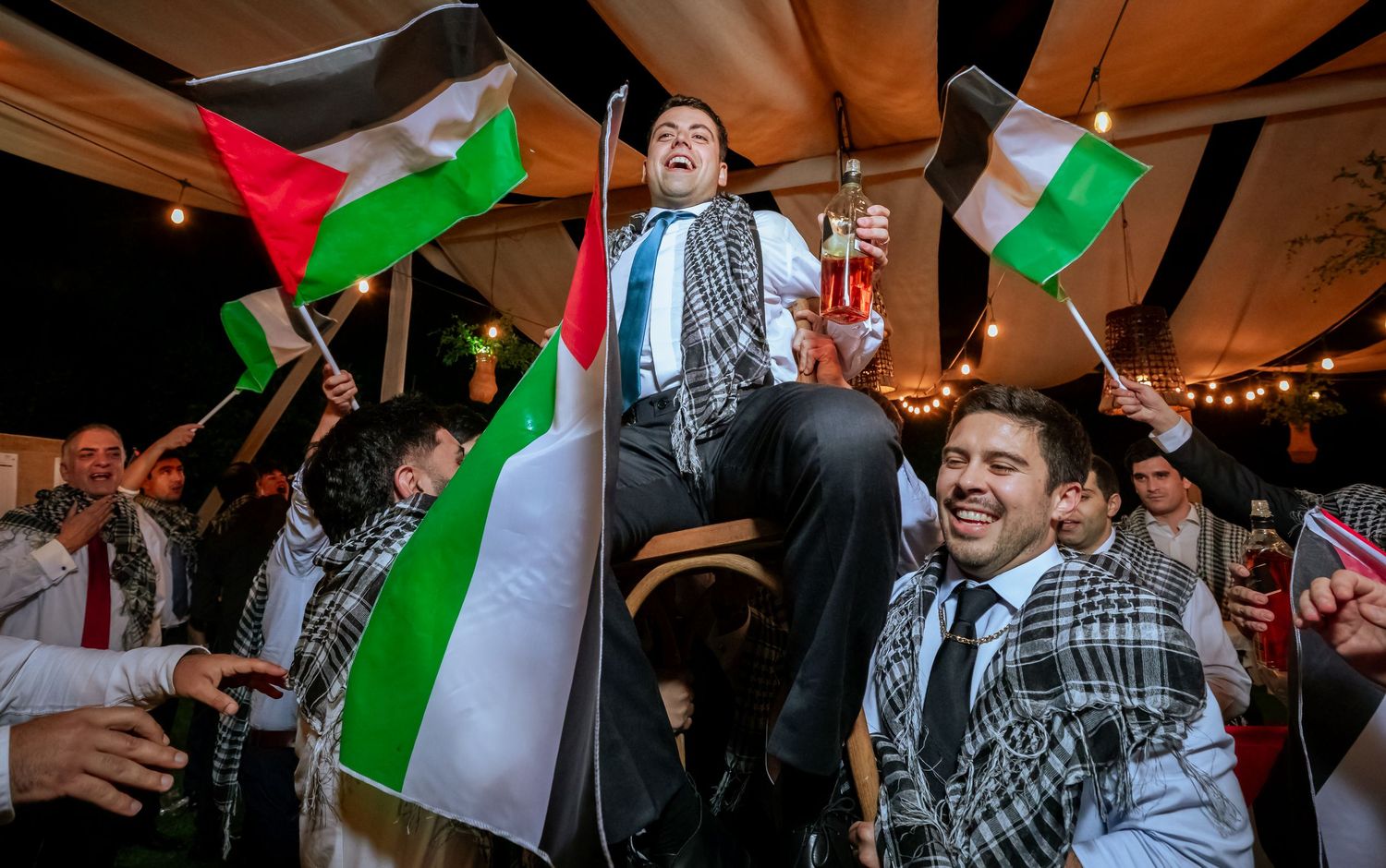Groom lifted by celebrating guests waving Palestinian flags at a festive wedding reception.