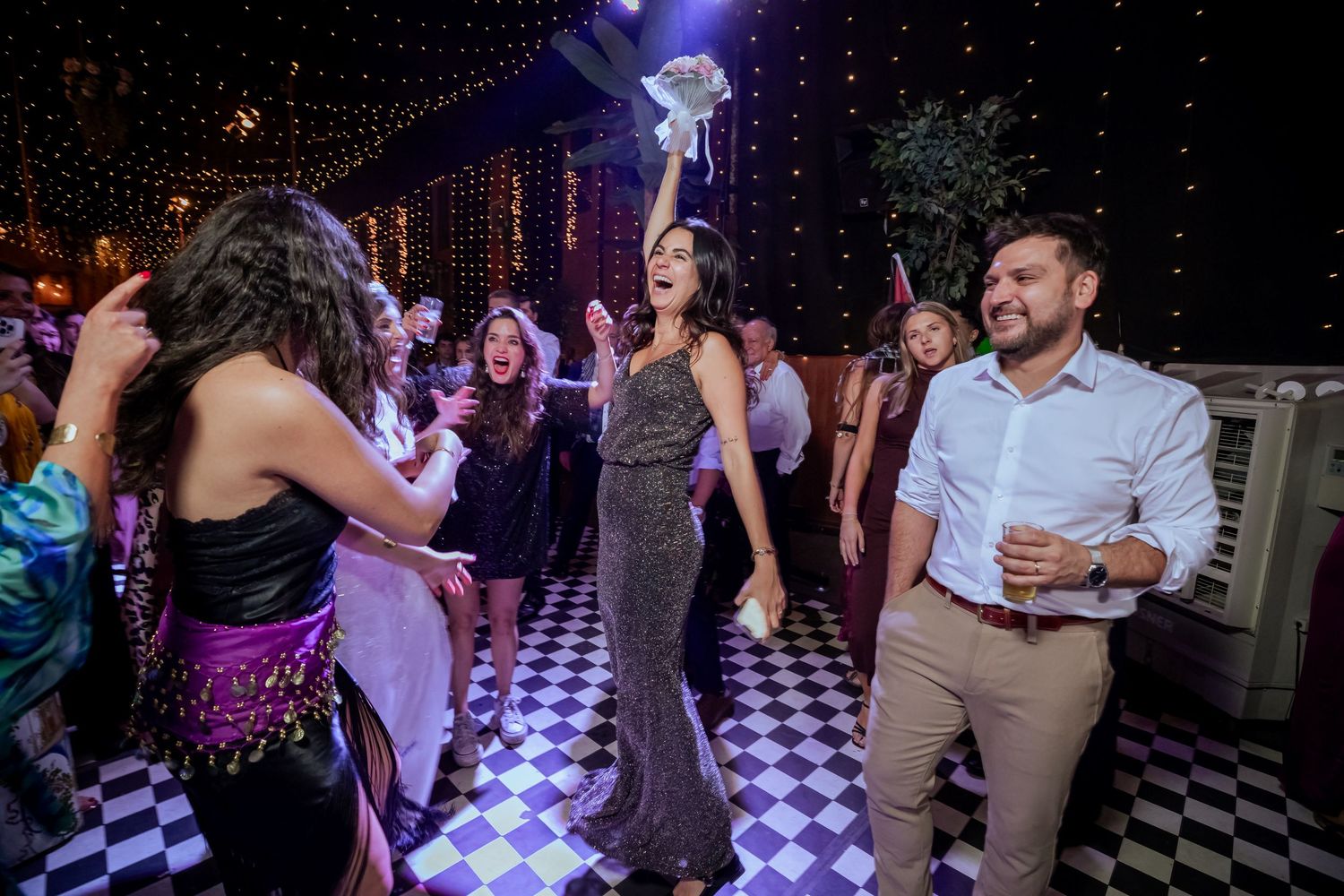 Bride in silver gown tosses bouquet to excited guests on checkered dance floor at wedding.