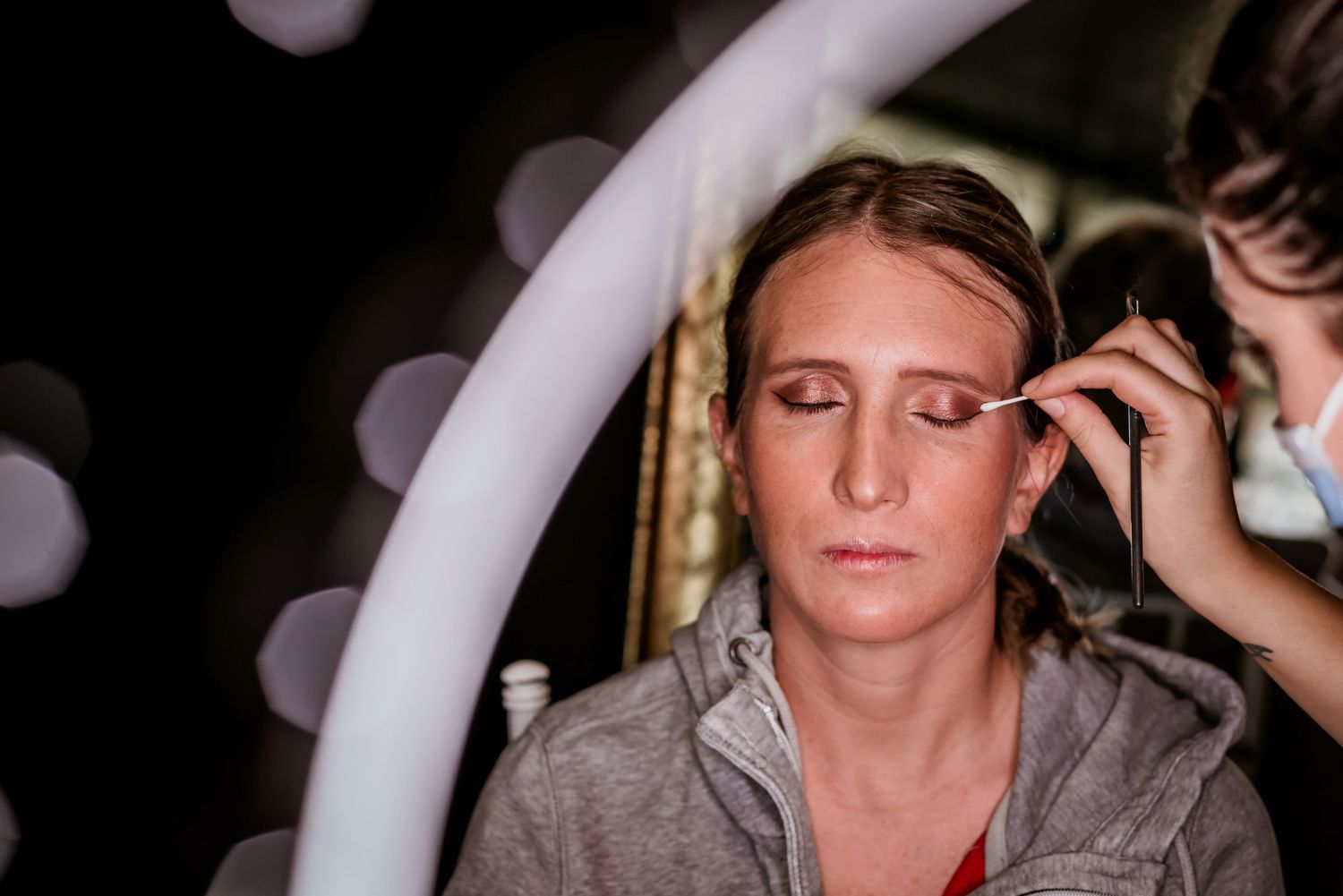 A makeup artist carefully applies eyeshadow to a woman wearing a gray hoodie.