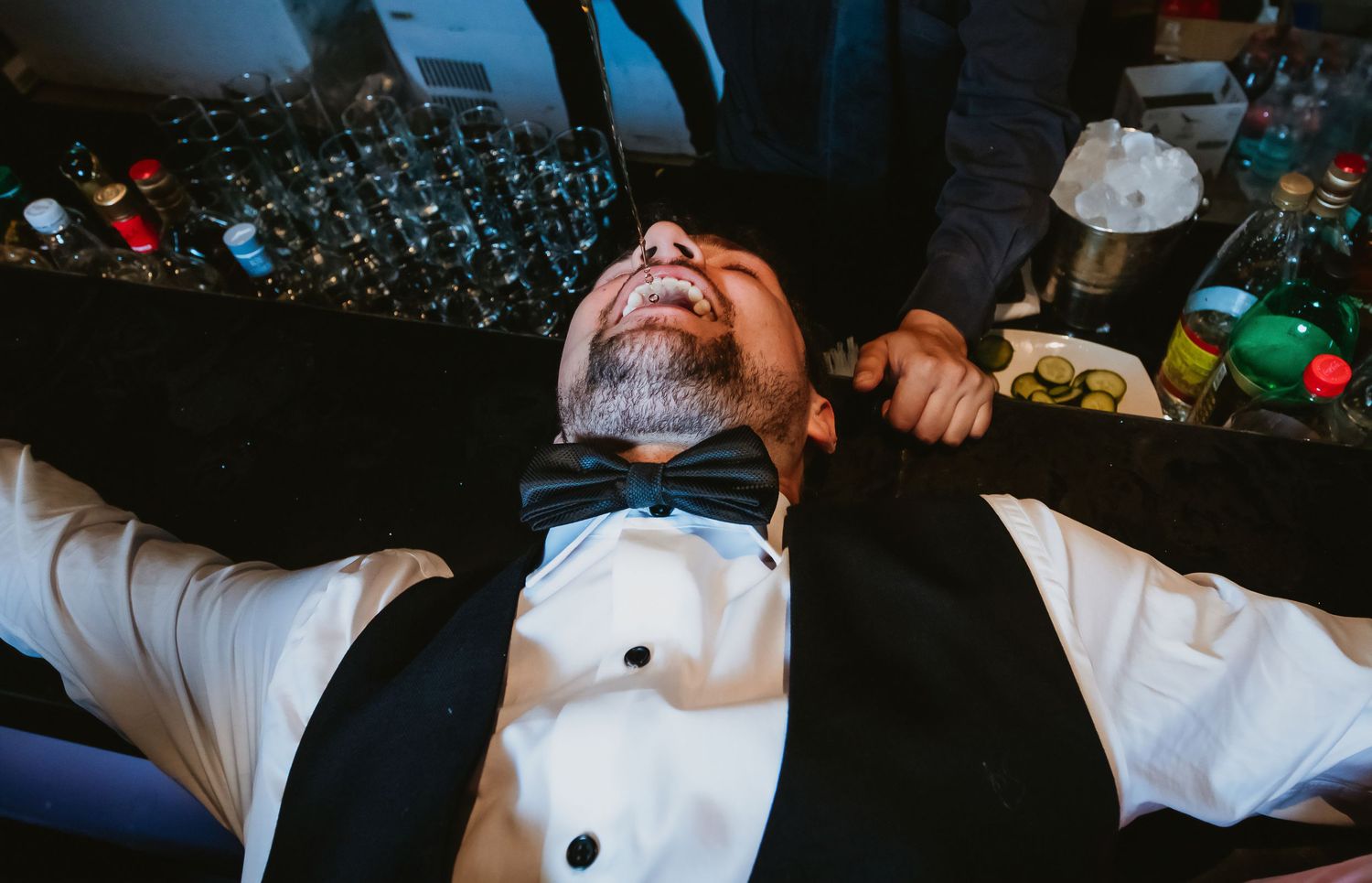 Man in tuxedo laughs while lying on bar surrounded by bottles at wedding reception.