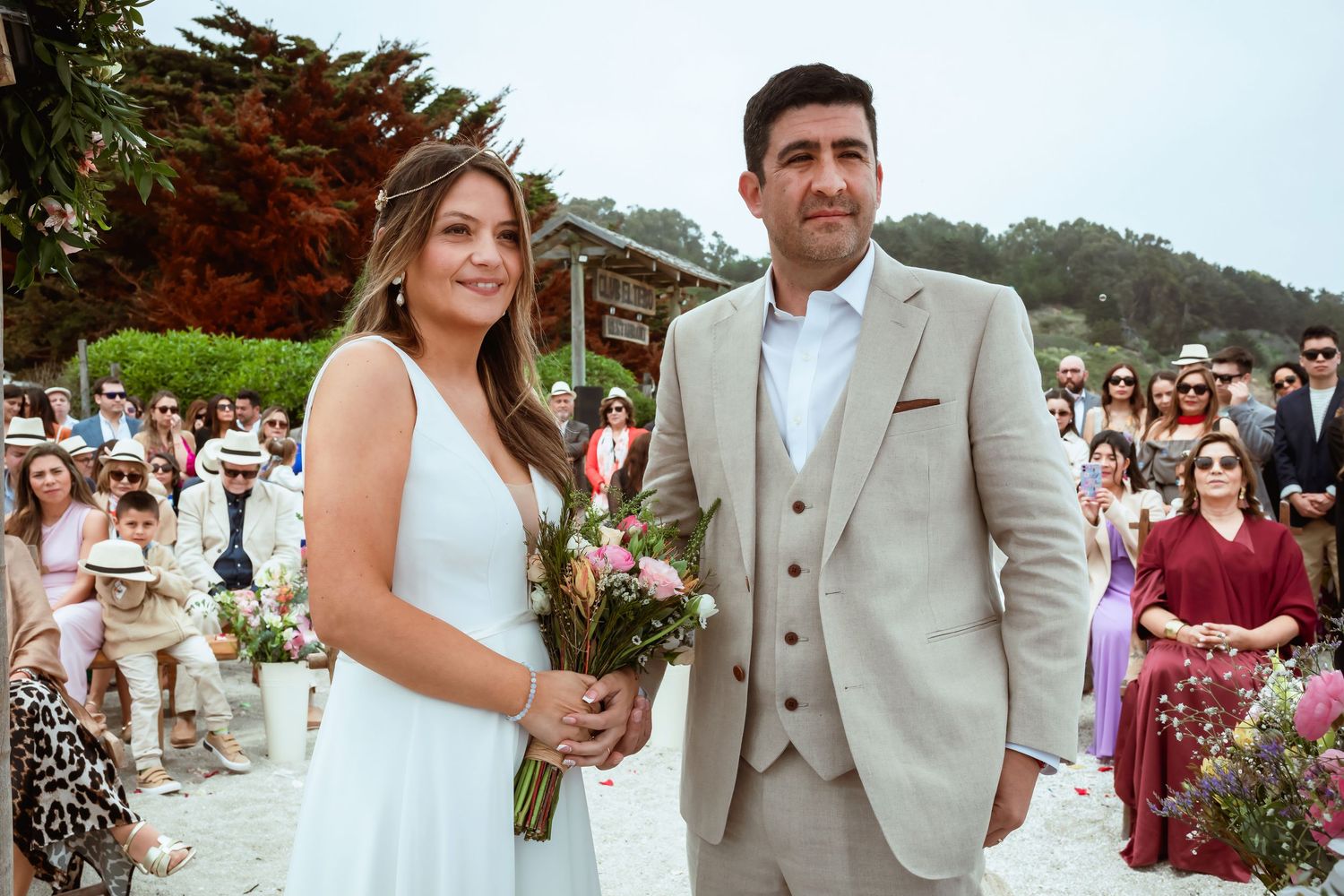 Newlywed couple poses together smiling after outdoor beach wedding ceremony with guests in background.