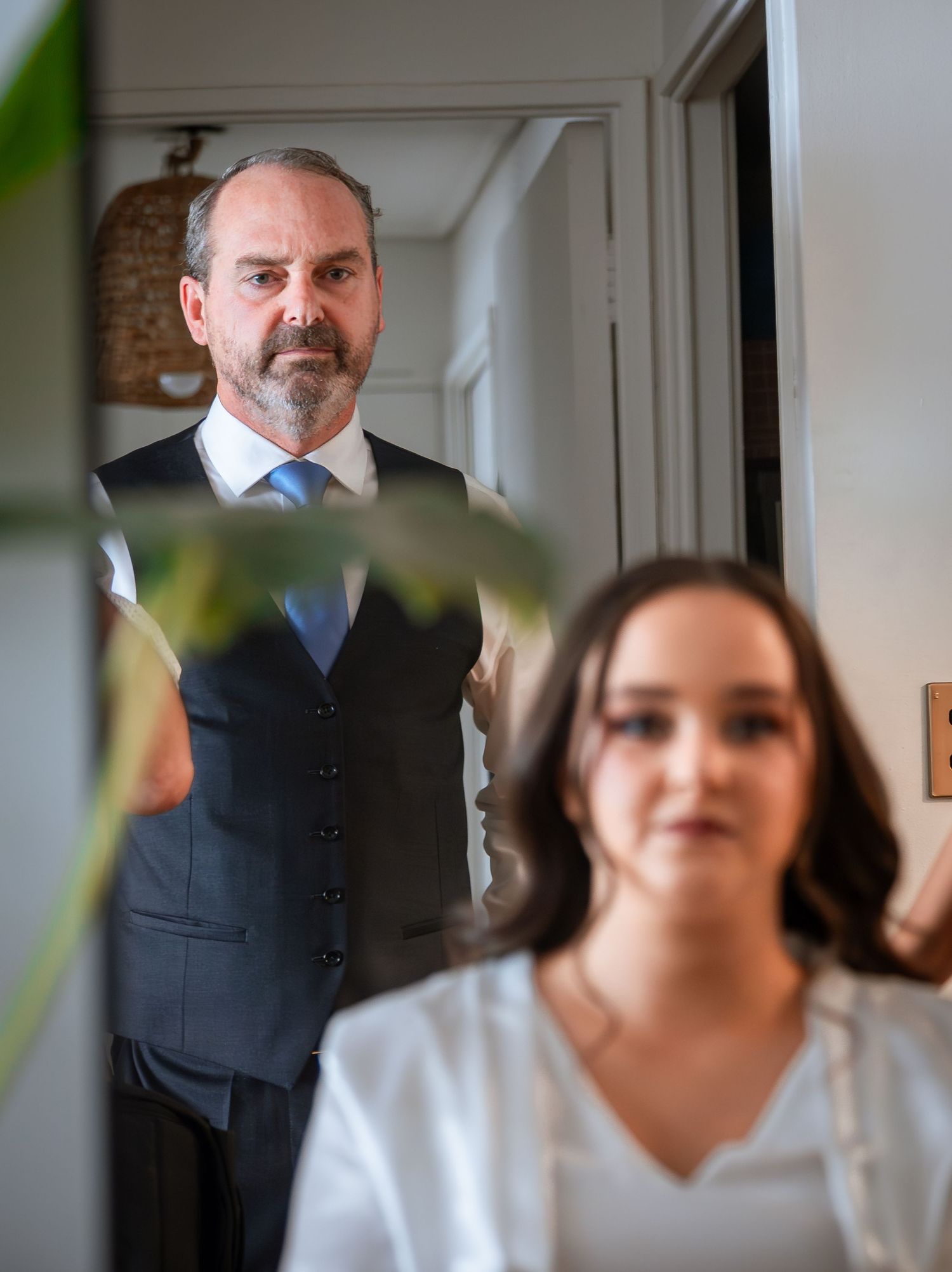 Groom in dark vest and tie stands behind bride during wedding preparations.