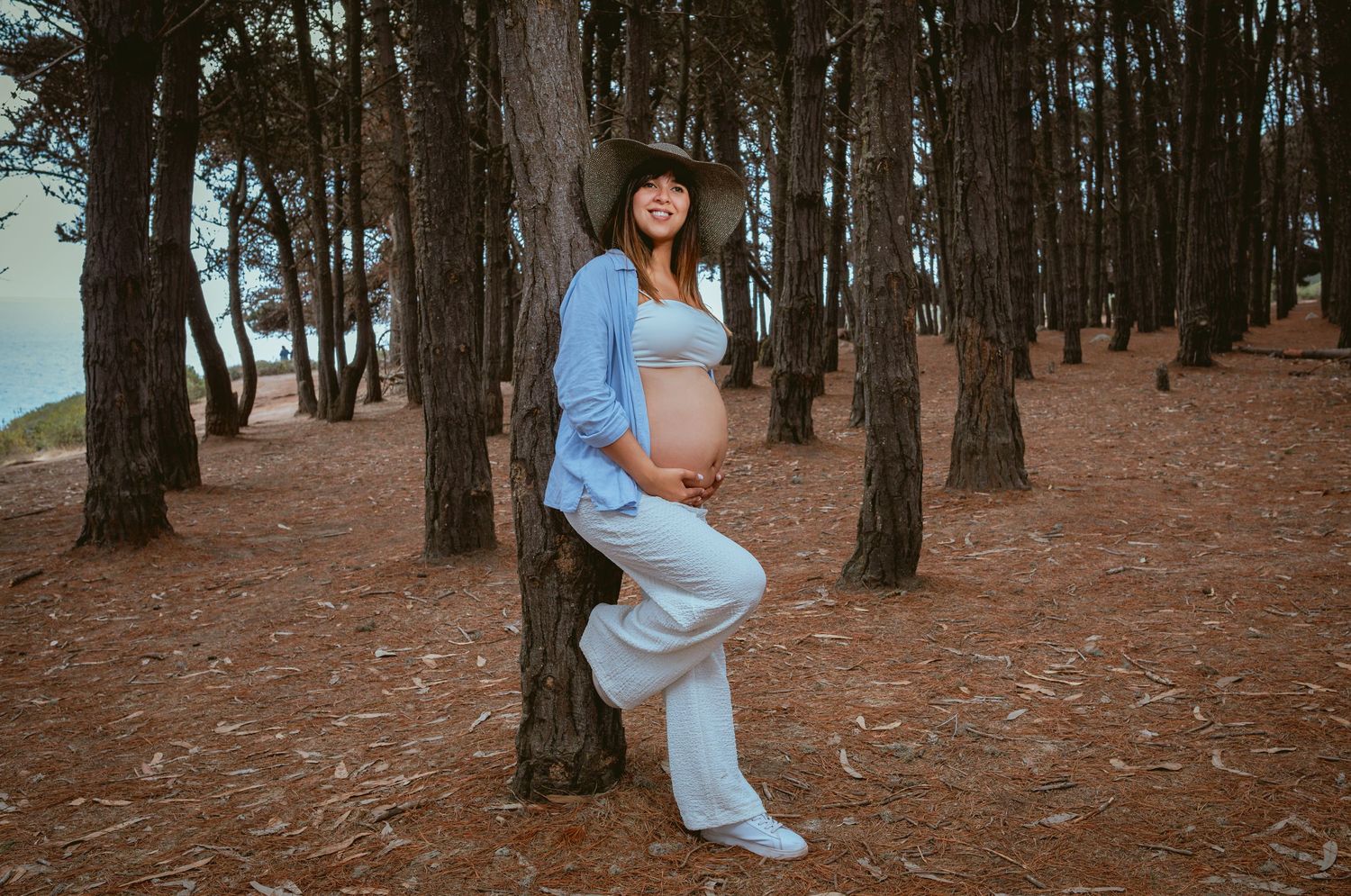 Pregnant woman in blue outfit leaning against pine tree wearing hat.