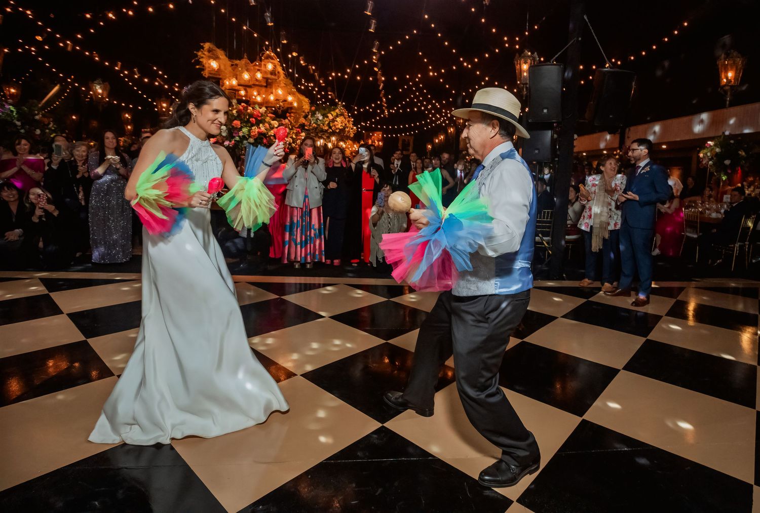 Bride and performer dance with colorful fans on checkered floor at festive wedding reception with guests watching.
