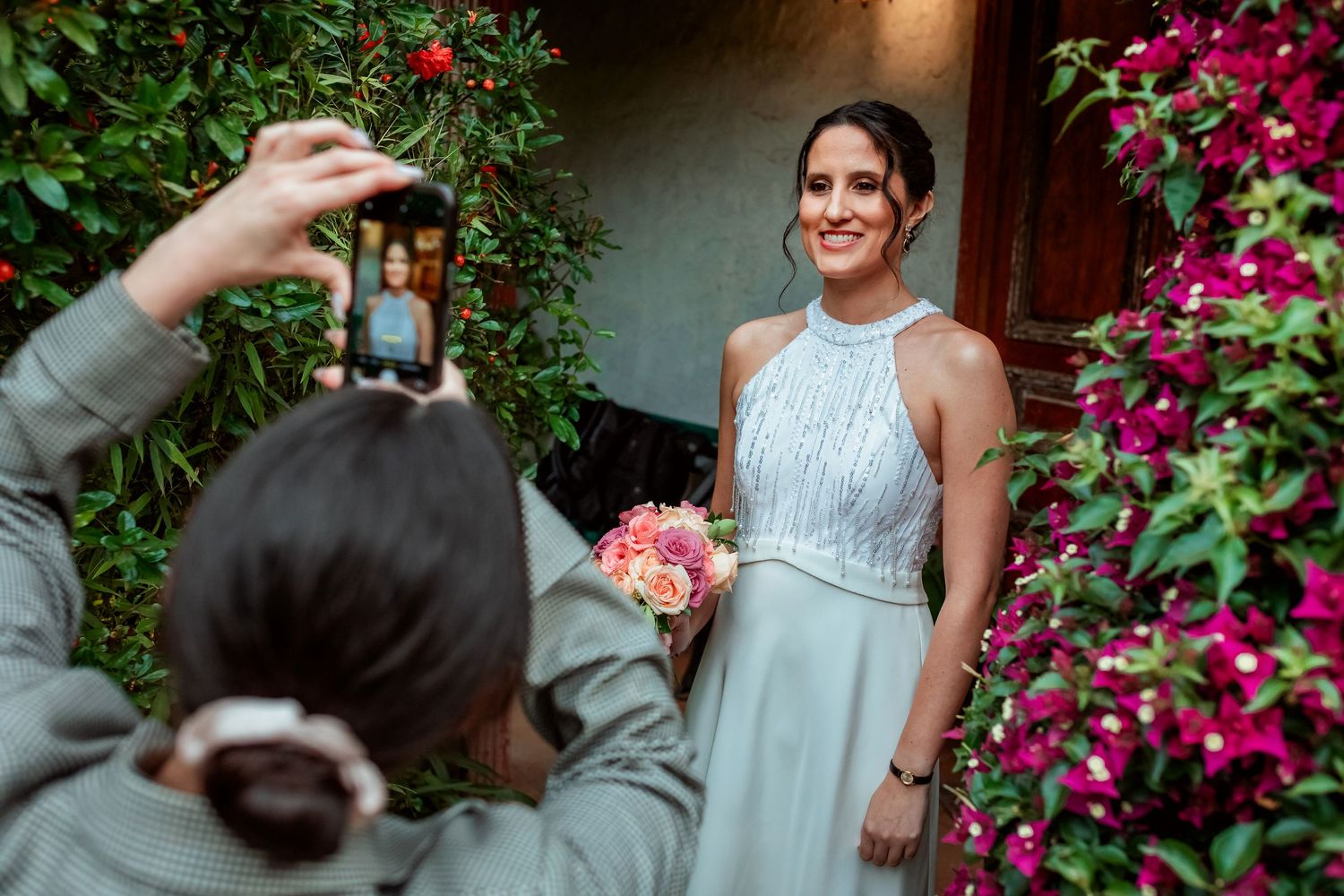 Bridesmaid photographs elegant bride holding pink bouquet in silver halter gown among vibrant flowering garden walls.