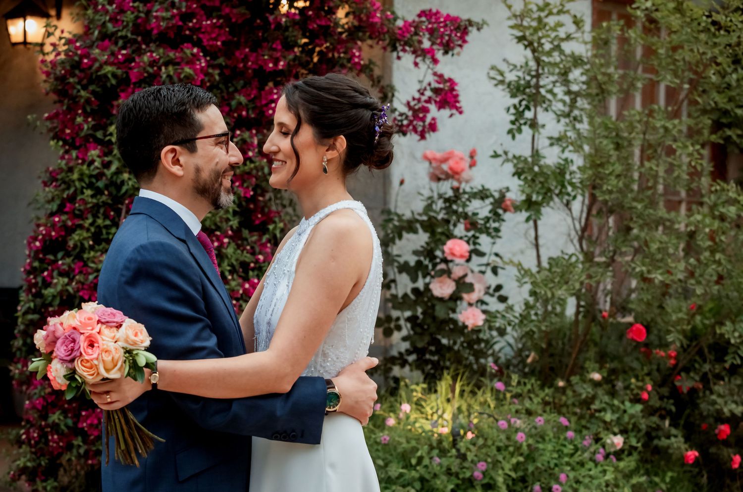Bride and groom sharing a romantic moment surrounded by vibrant pink bougainvillea and rose garden.