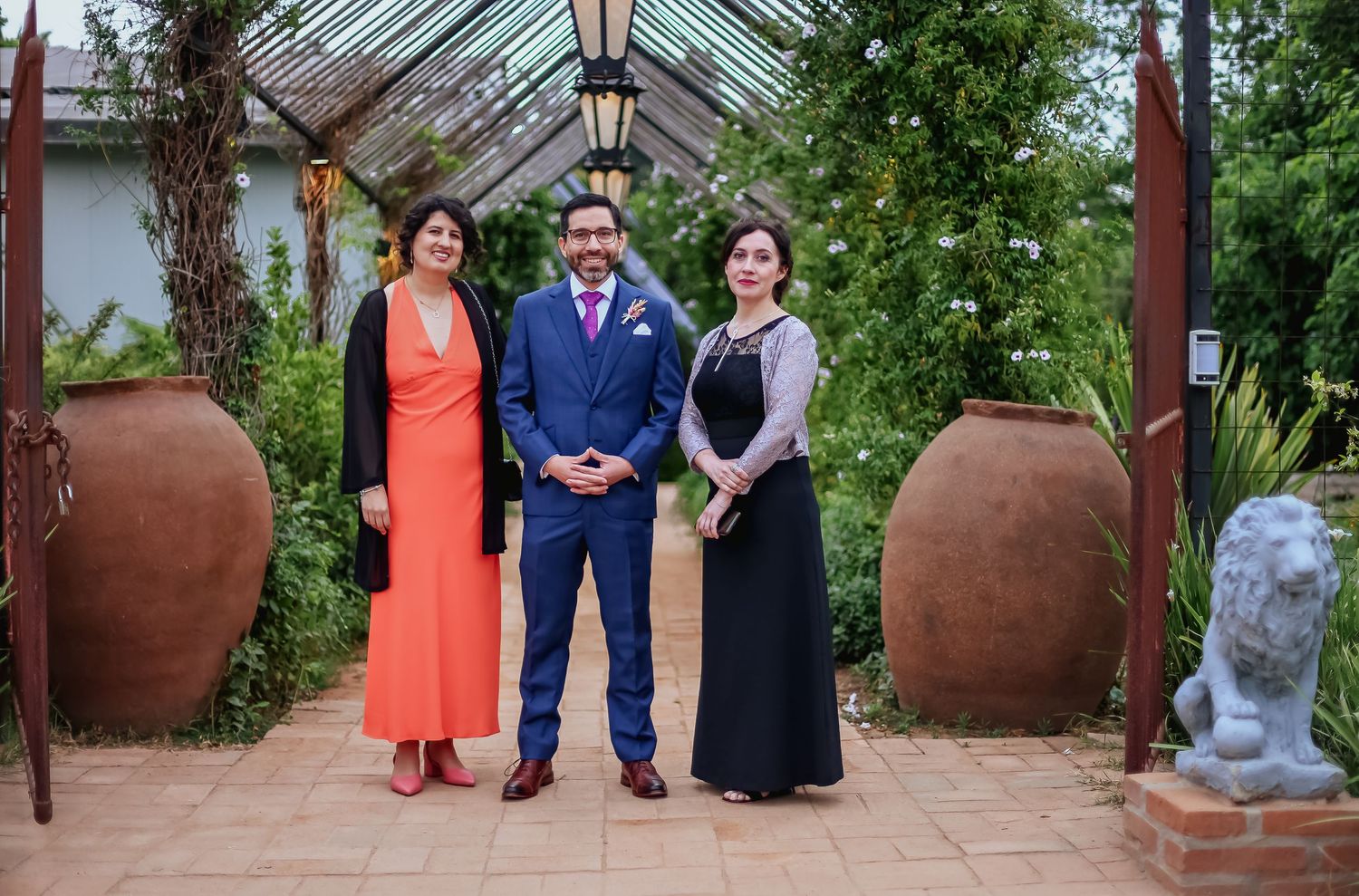 Groom in blue suit posing with two women in orange and black dresses in garden setting.