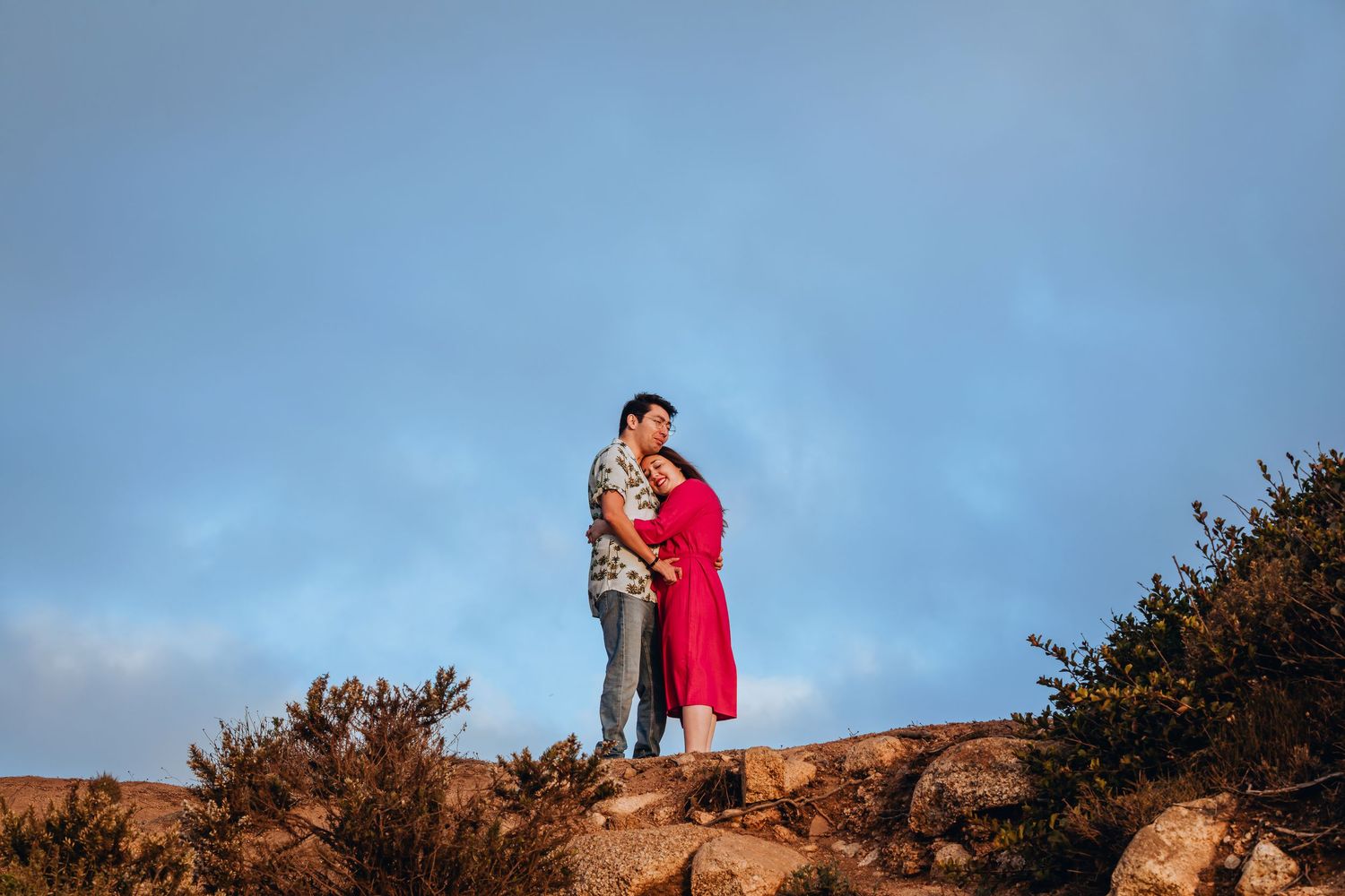 Couple embracing on rocky hilltop at sunset, woman in red dress and man in floral shirt under blue sky.
