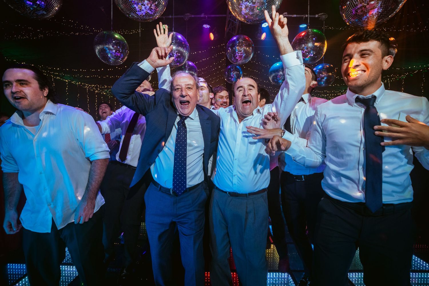 Men in dress shirts celebrating joyfully at a disco-themed party with mirror ball lights in the background.