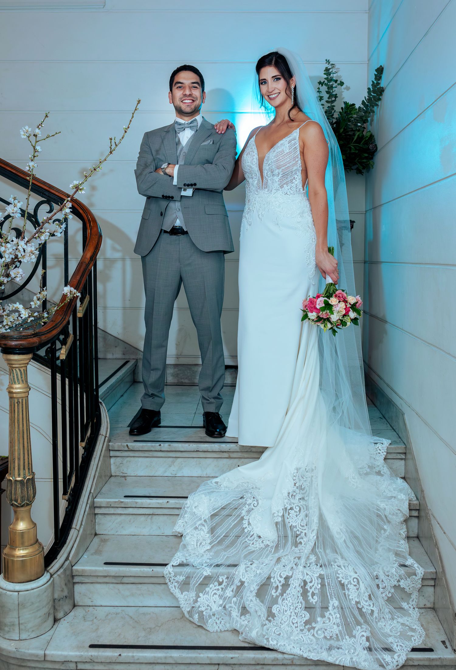 A bride in a lace wedding gown holding a bouquet poses with a groom in a gray suit on an elegant staircase.