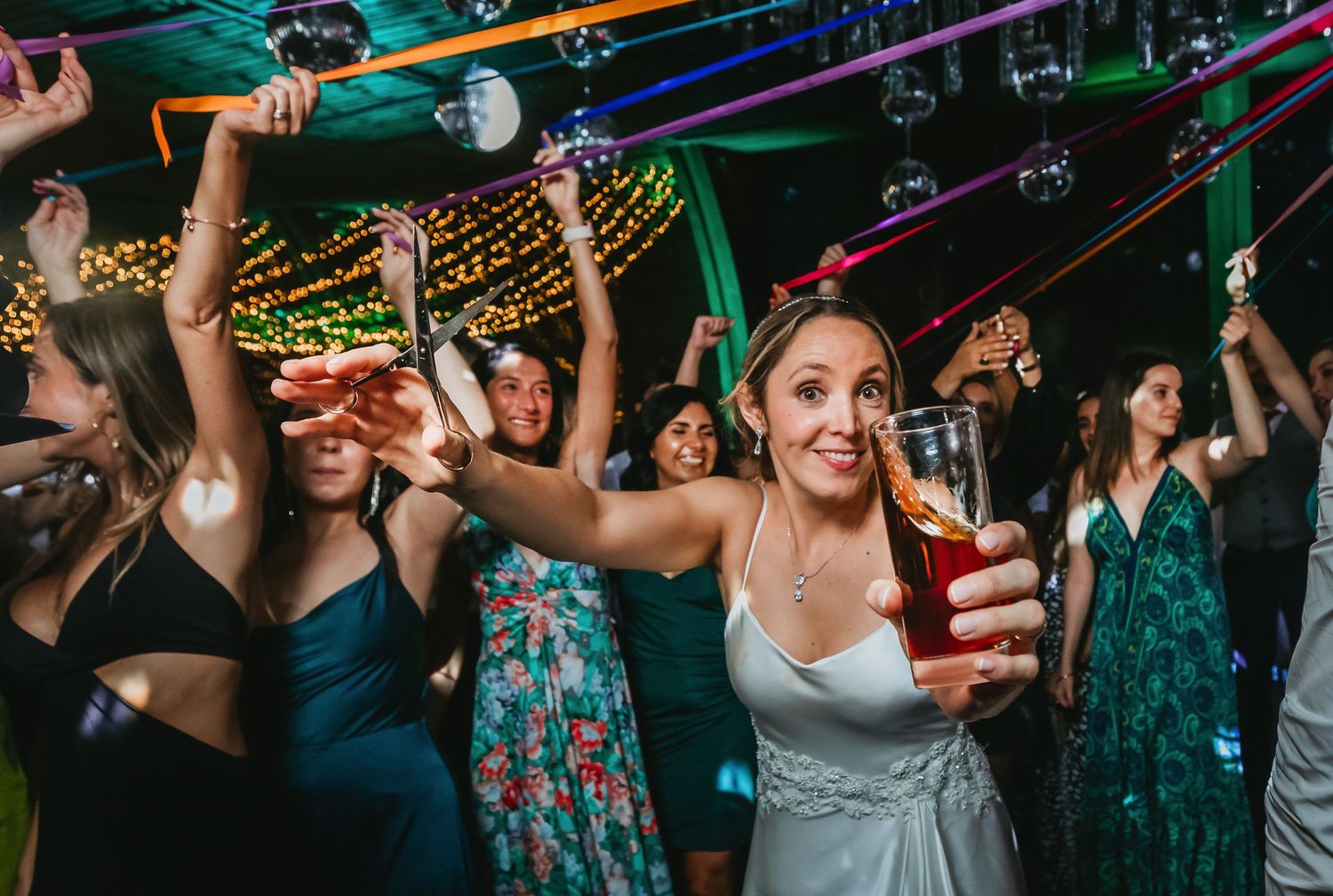 Smiling bride holding beer glass while dancing with guests at wedding.