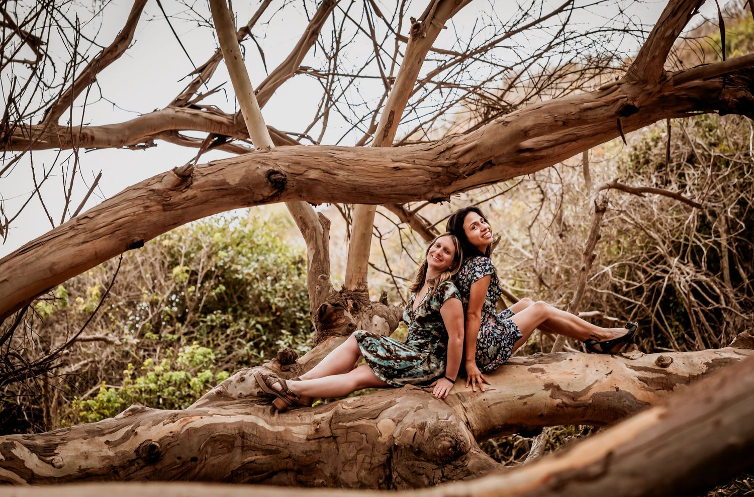Two women in matching floral dresses sitting together on a large driftwood log outdoors.