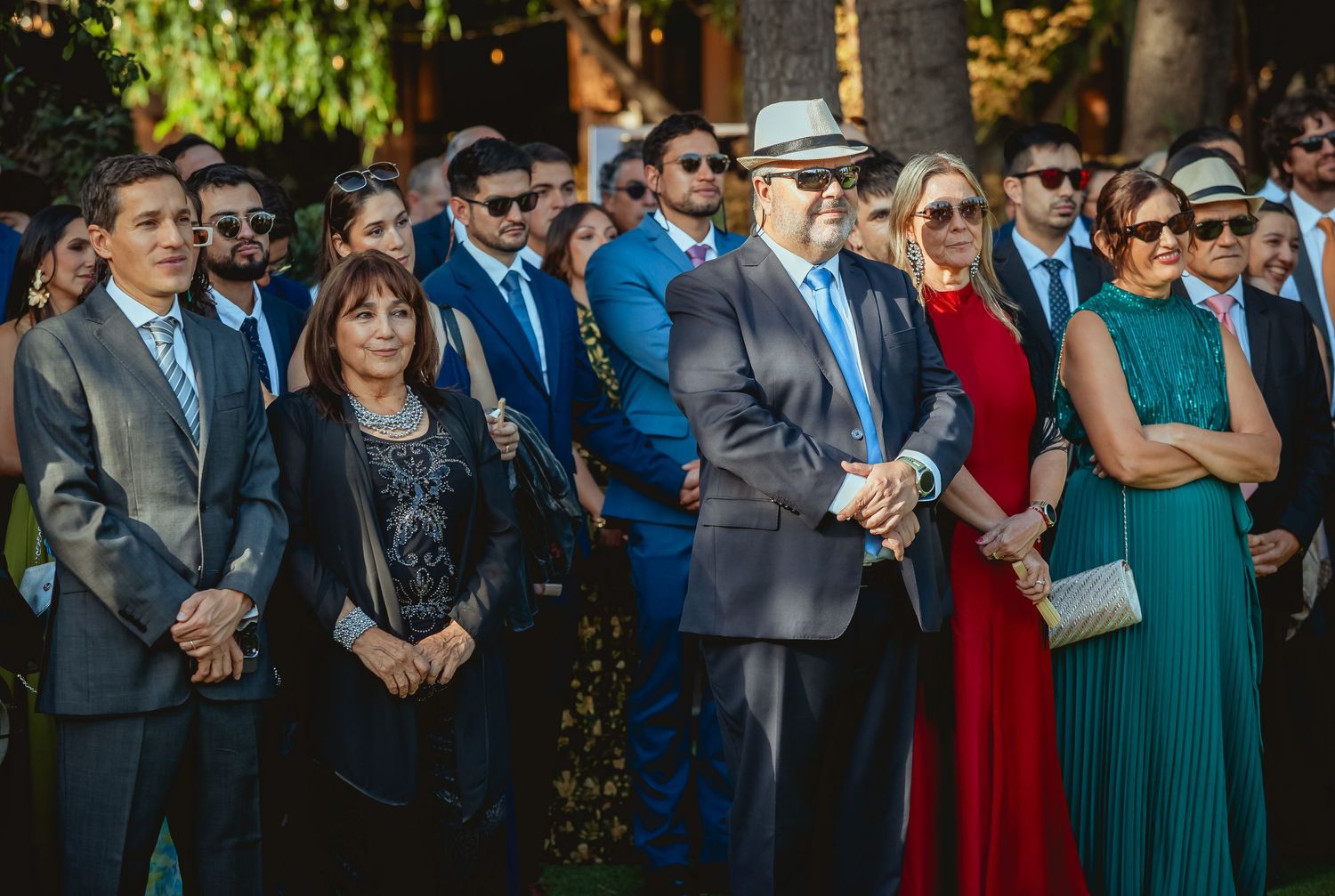 Group of well-dressed wedding guests stand together watching outdoor ceremony in garden setting.