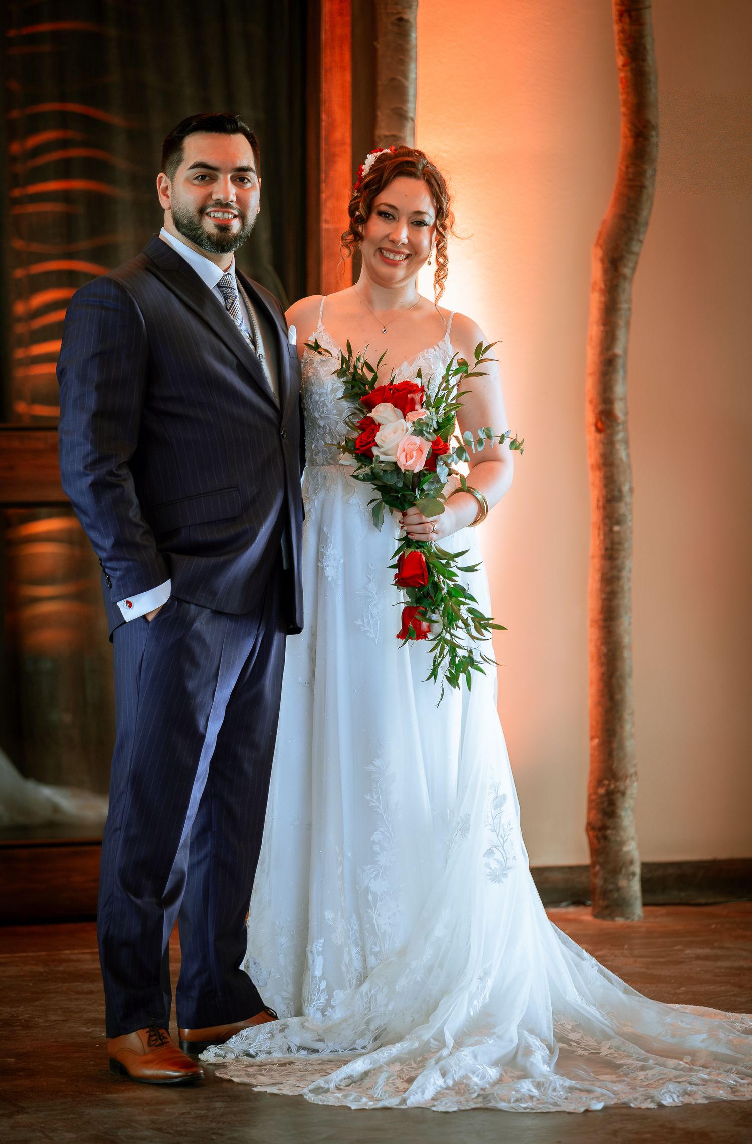 Bride holding a cascading red and pink rose bouquet poses with groom in warm wedding venue lighting.