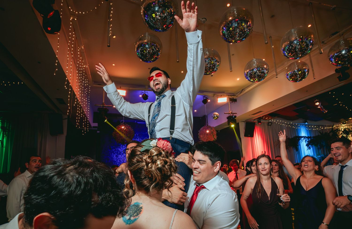 Groom with red nose lifted by cheering guests on a vibrant wedding dance floor.