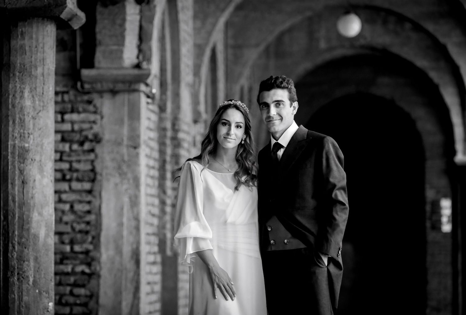 Black and white portrait of bride and groom under brick archways at historic venue.