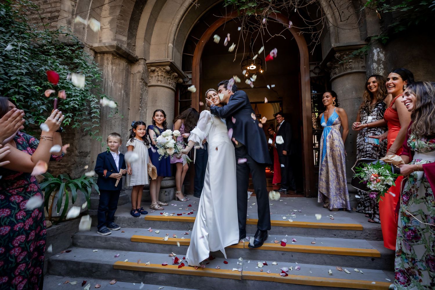 Newlyweds share a romantic kiss on church steps surrounded by petal-throwing guests.