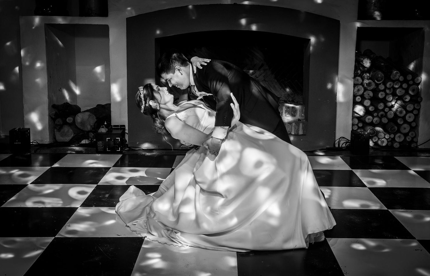 Black and white photo of bride and groom sharing intimate dip kiss on checkered dance floor.