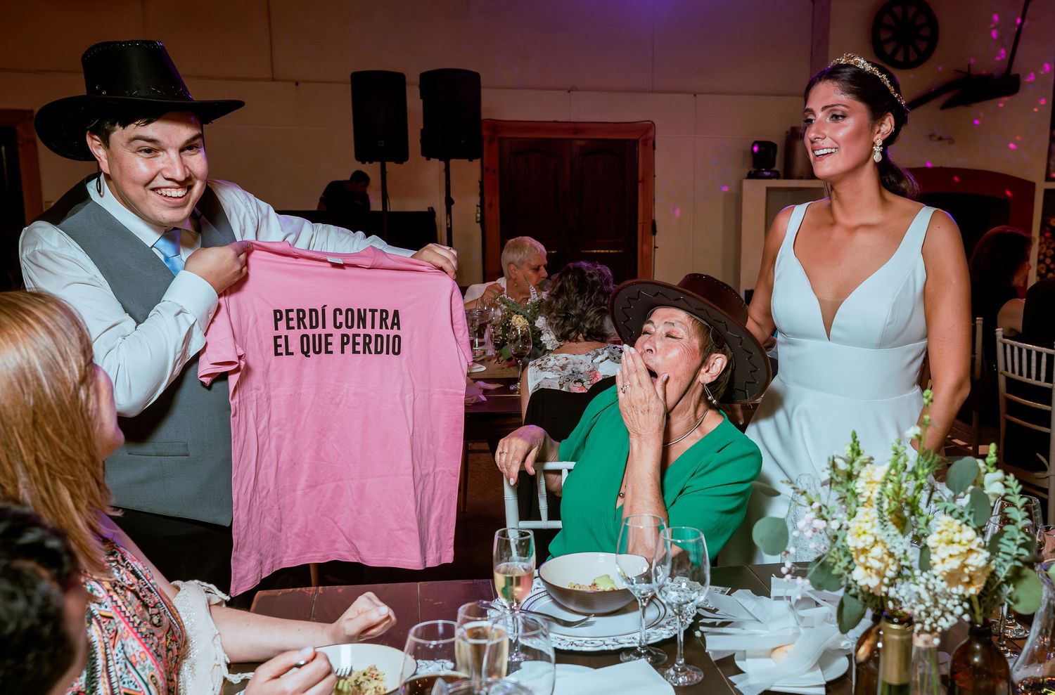 Groom holds pink Spanish shirt while bride laughs at wedding reception dinner table.