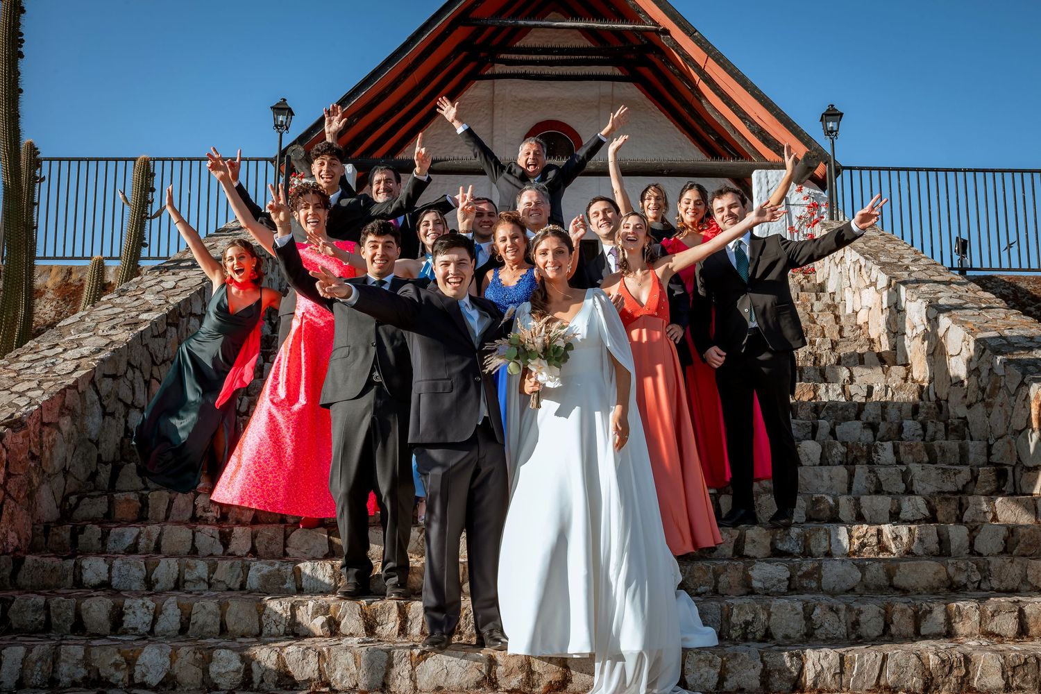 Joyful wedding party celebrating on stone steps outside rustic chapel with raised arms.