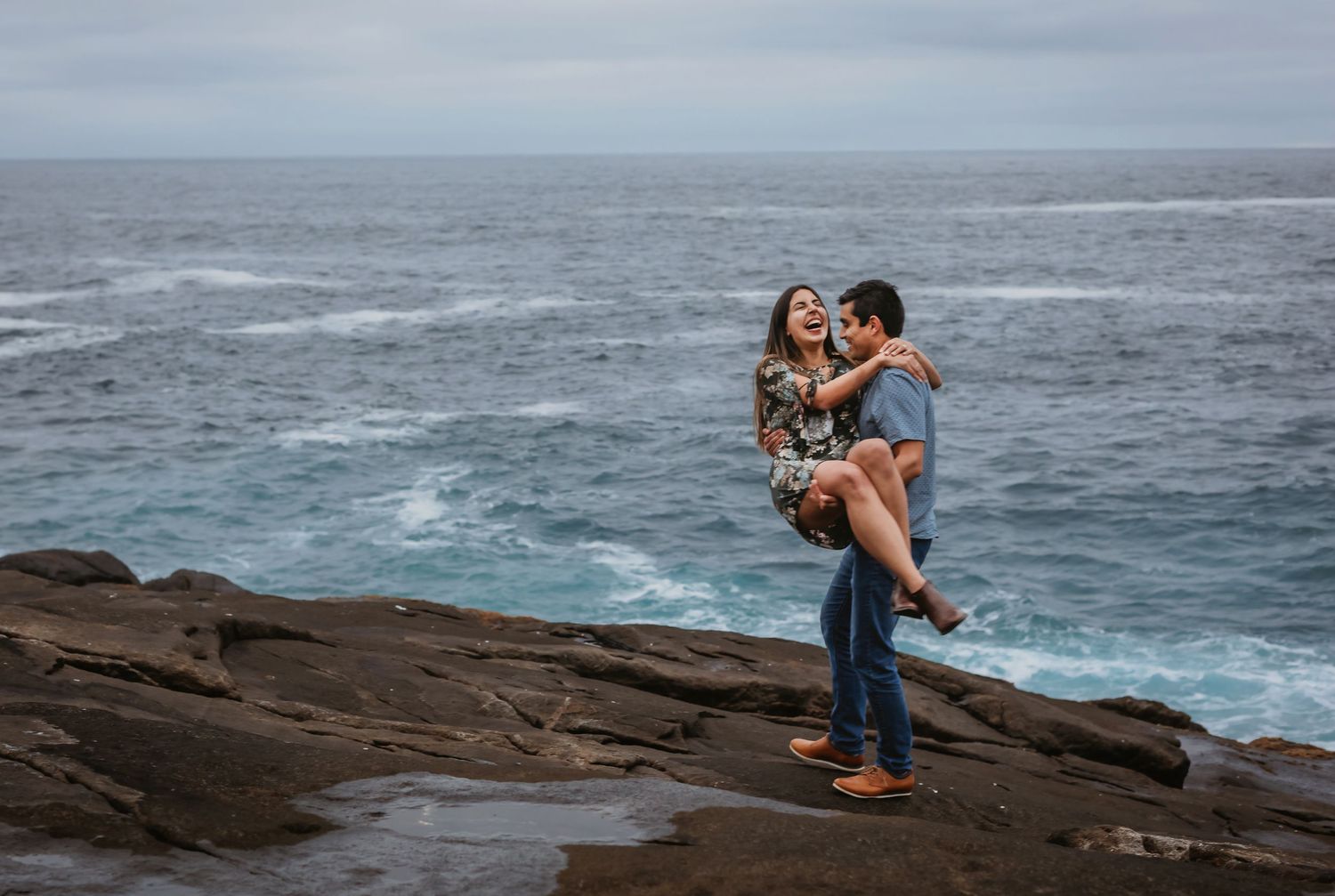 Couple sharing a romantic moment on rocky coastal cliffs with dramatic ocean waves in the background.