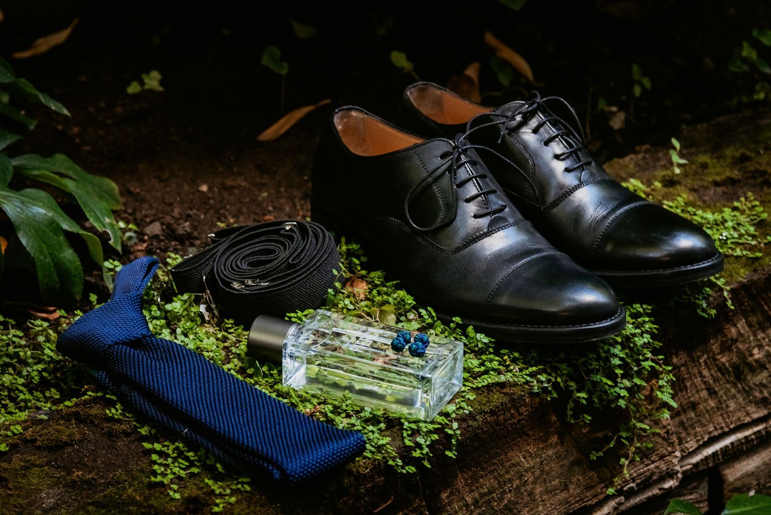Black dress shoes, blue tie, belt, and cologne arranged on mossy surface outdoors.