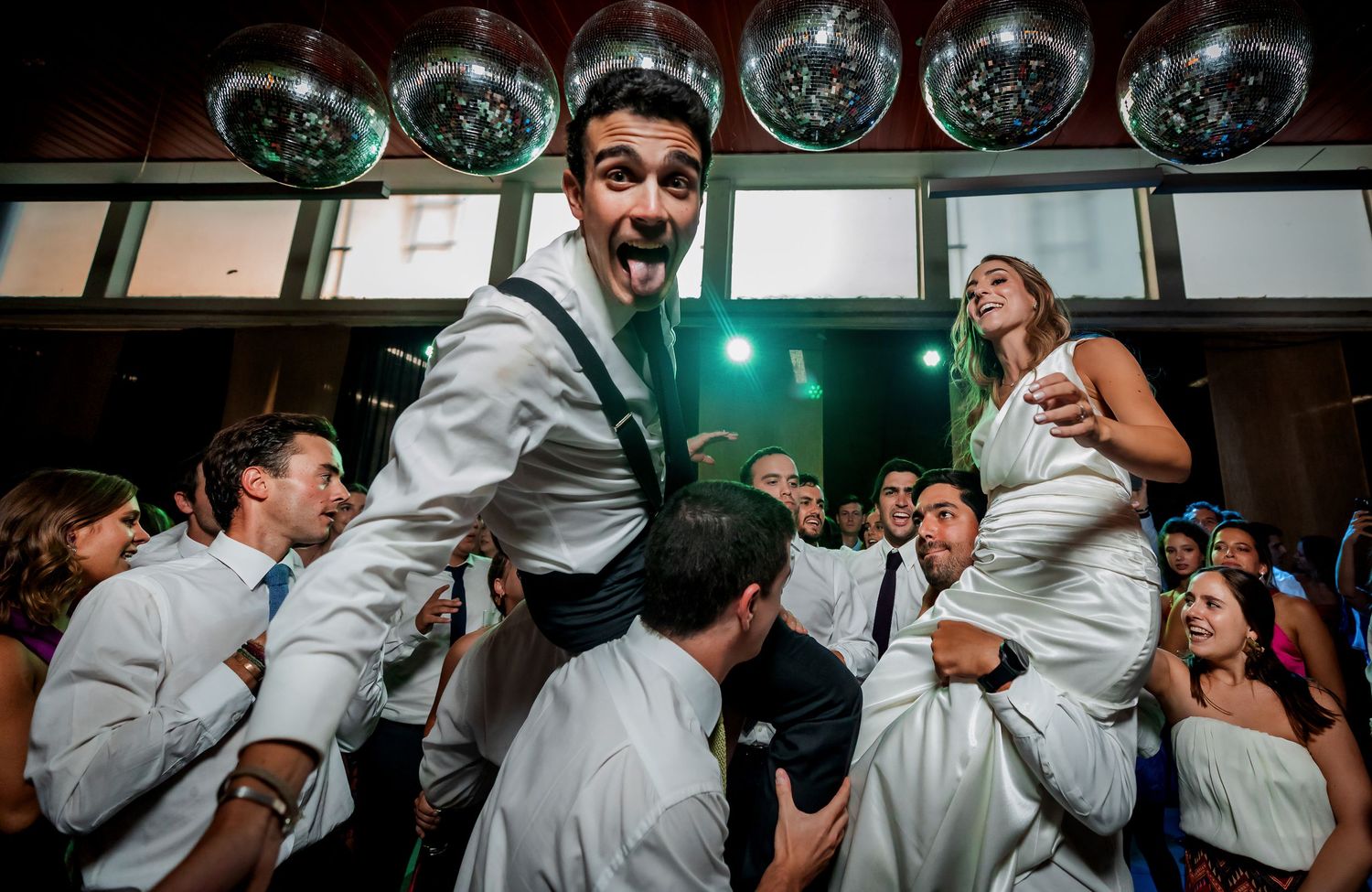Exuberant groom crowd-surfing with open mouth expression under disco balls at lively wedding reception party.