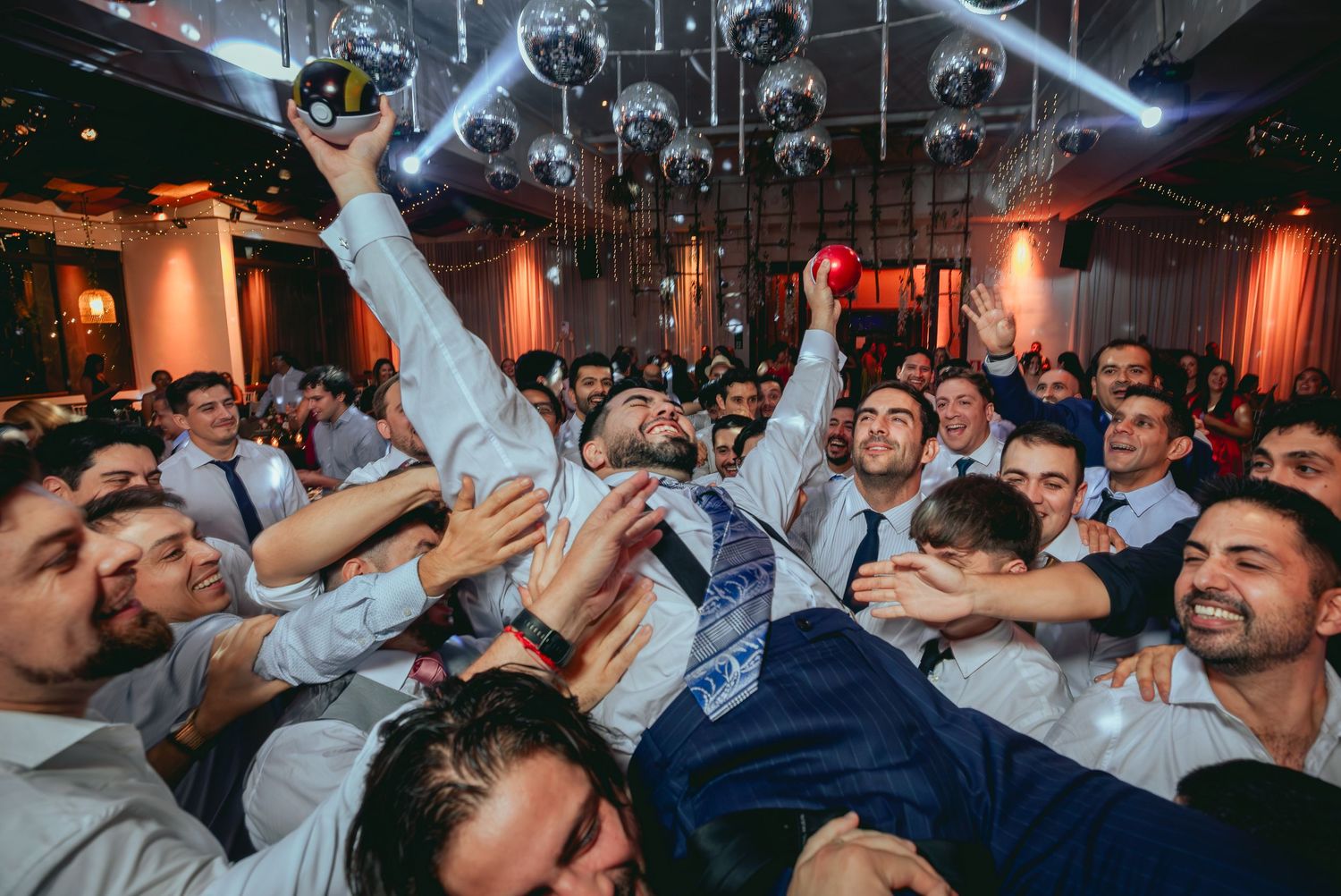 Groom lifted by cheering groomsmen in celebratory crowd surf at wedding reception.