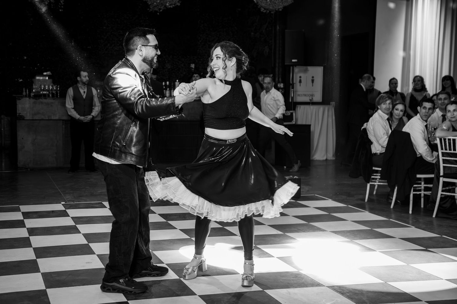 Couple performs energetic dance on black and white checkered floor at wedding reception.