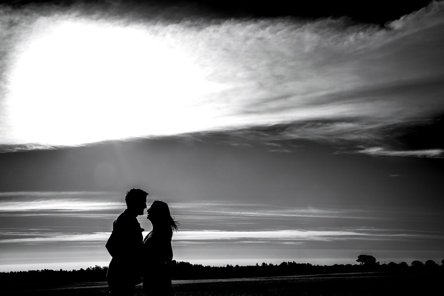 Romantic black and white silhouette of a couple about to kiss against a bright cloudy sky.