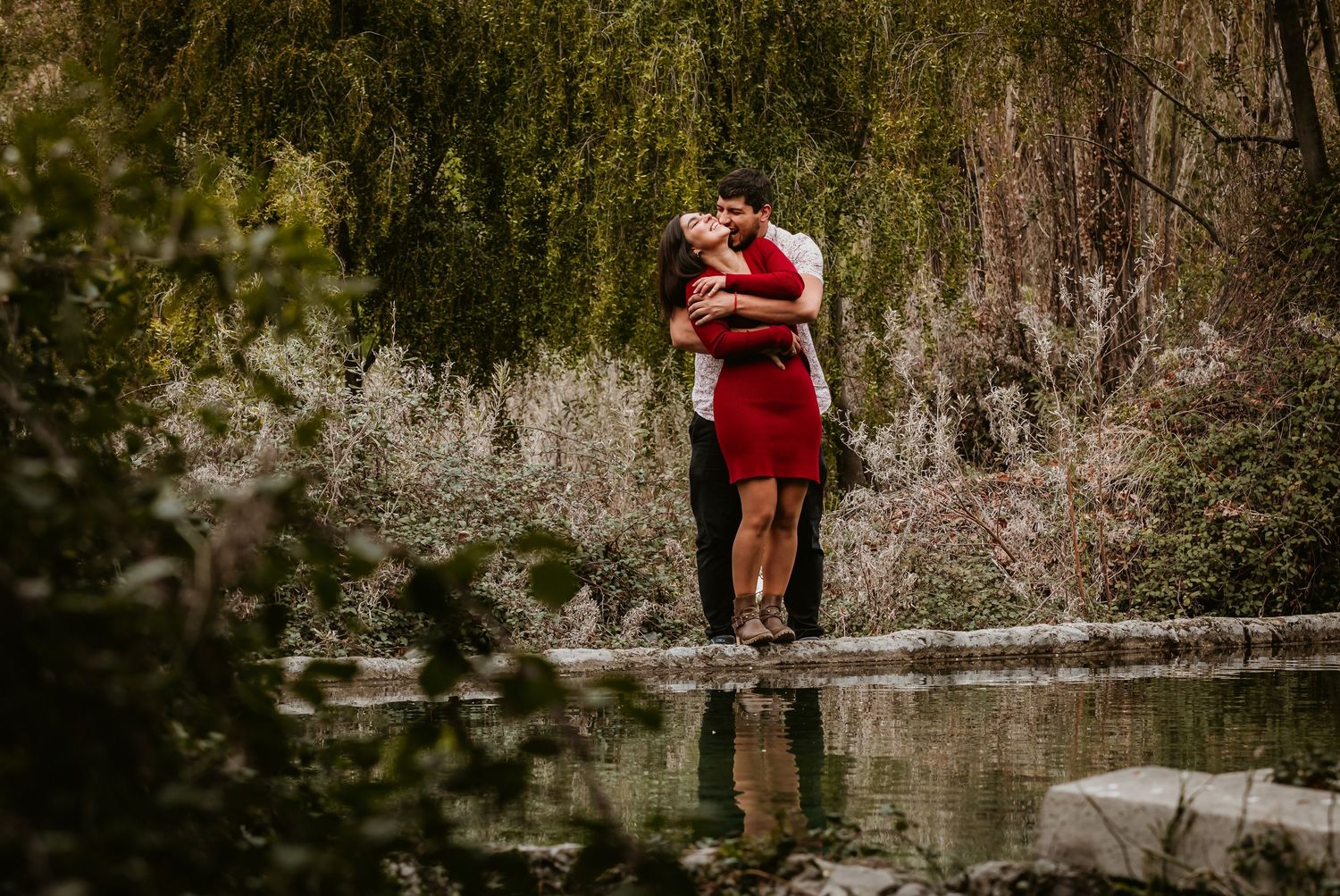 Couple sharing a kiss near a serene pond surrounded by lush green trees.