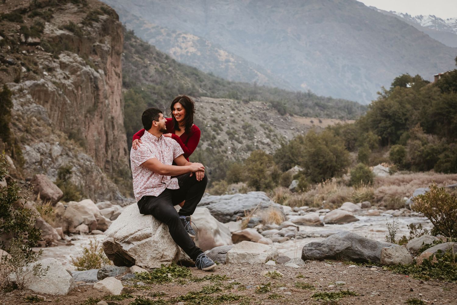 Couple sitting together on rocks with a scenic mountain river landscape in the background.