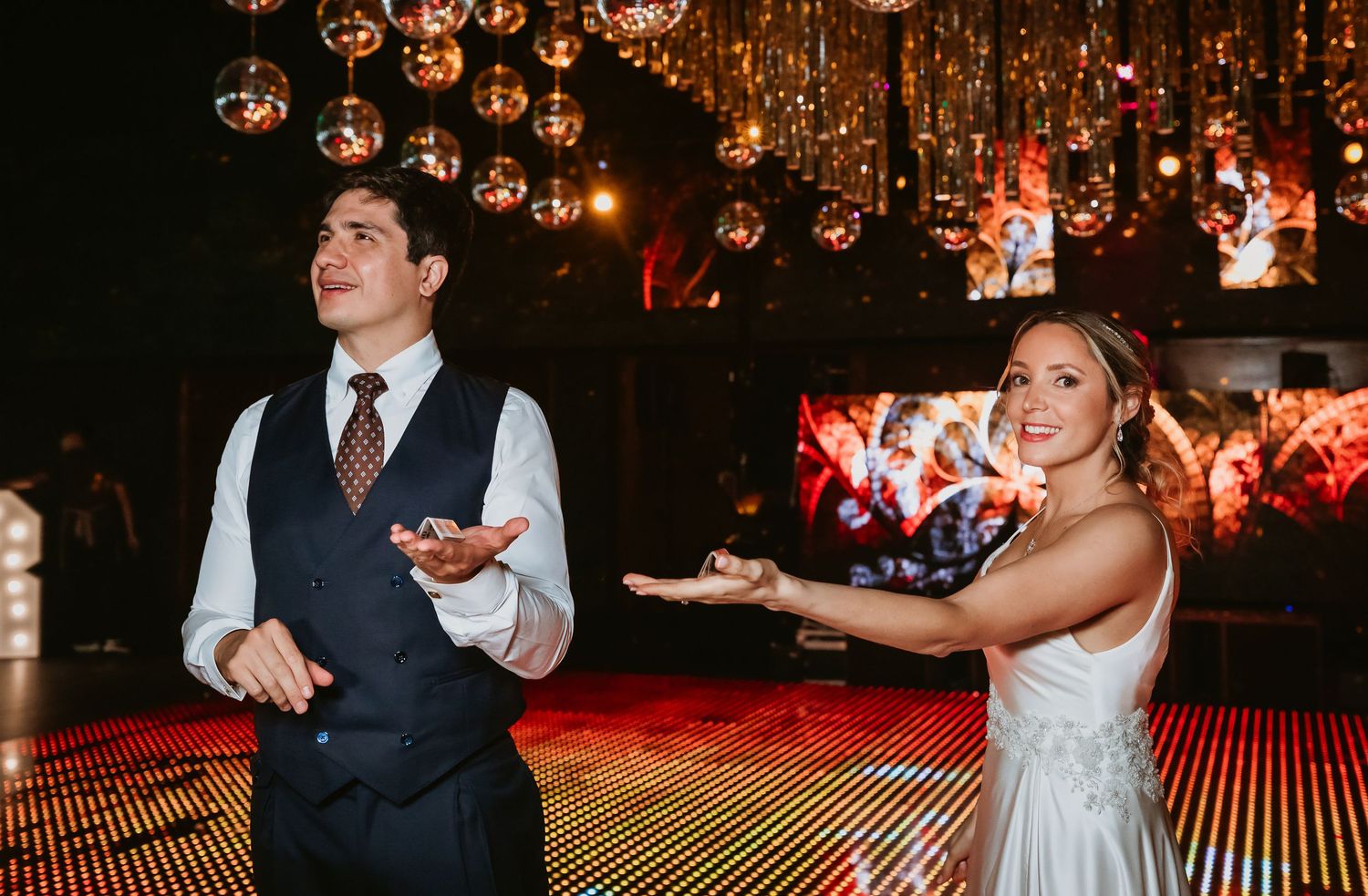 Bride and magician performing a card trick together on a sparkling stage at a wedding reception.