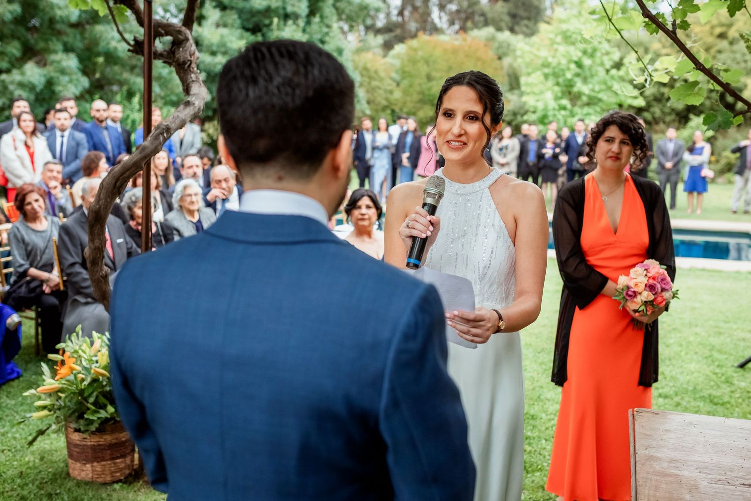 Bride speaks into microphone during outdoor wedding ceremony with guests seated behind.