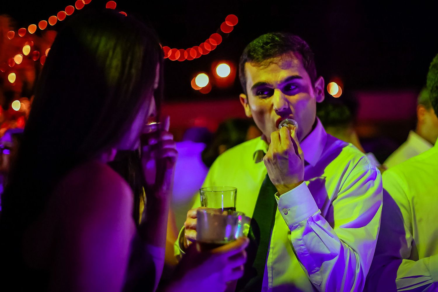Man in white dress shirt smokes a cigar while holding a drink at an outdoor evening party.