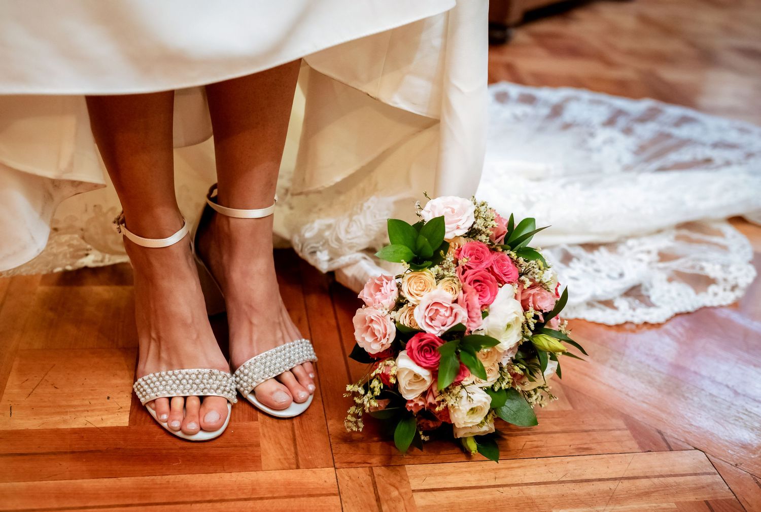 Bride wearing silver ankle-strap heels standing beside a pink and white rose wedding bouquet on hardwood floor.