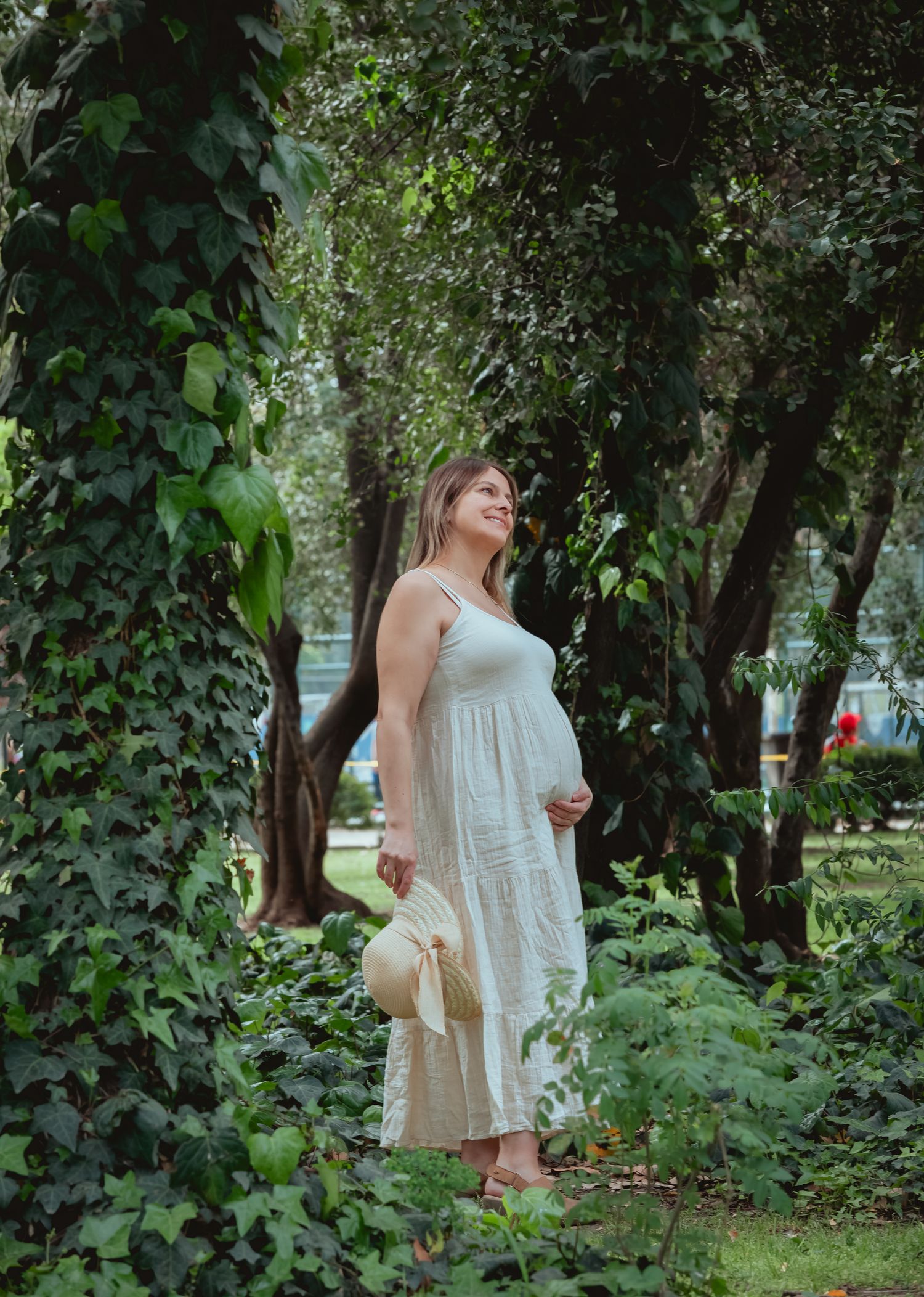 Pregnant woman in white maxi dress holding straw hat while standing among ivy-covered trees outdoors.