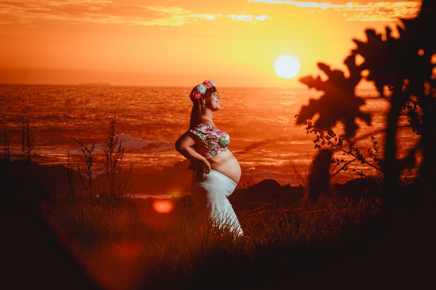 Pregnant woman in white gown and flower crown gazes upward during a radiant red sunset.